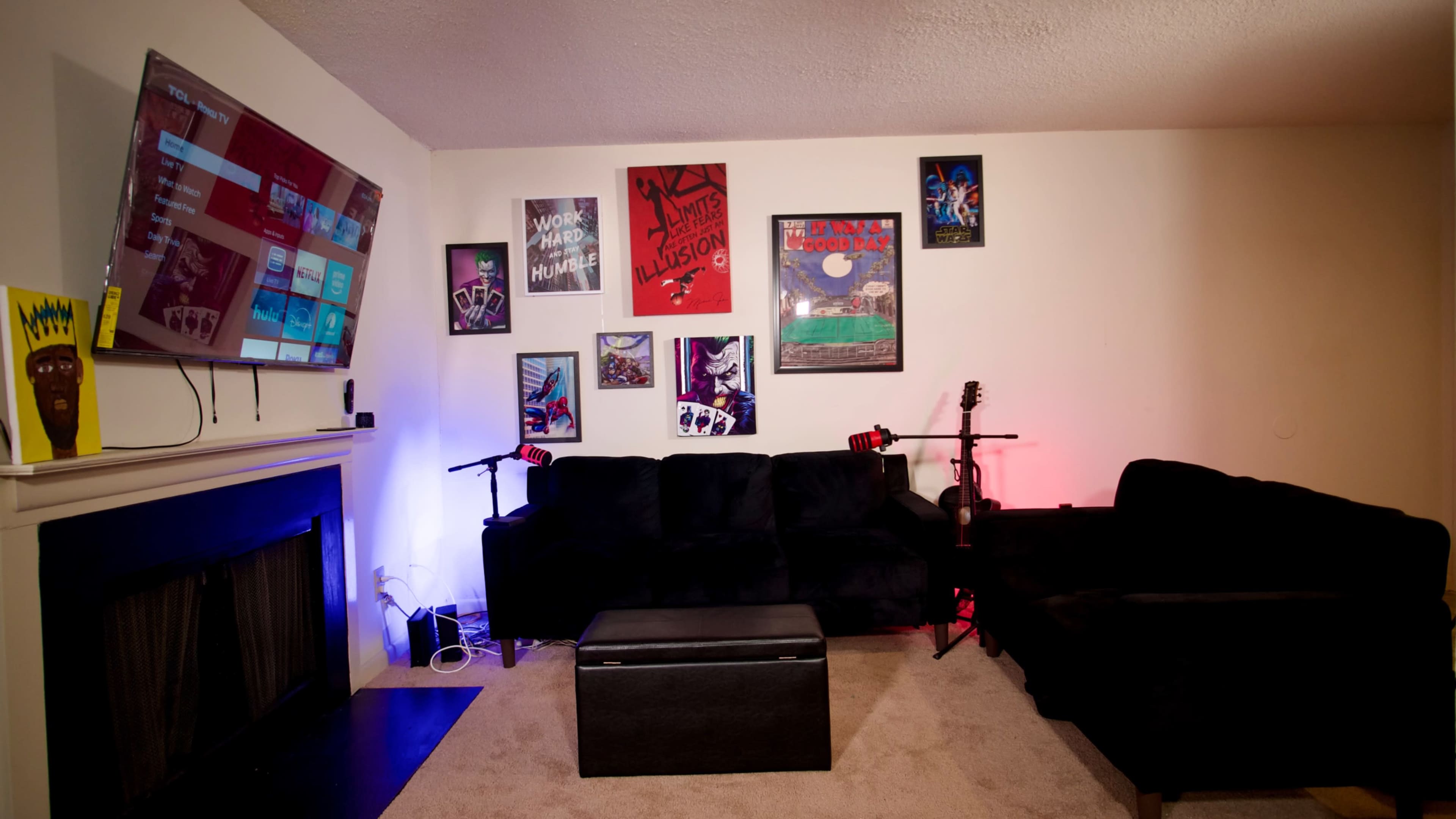 A living room features a black sectional sofa, a coffee table, and a wall adorned with various posters, while a television displays a screen in the background.