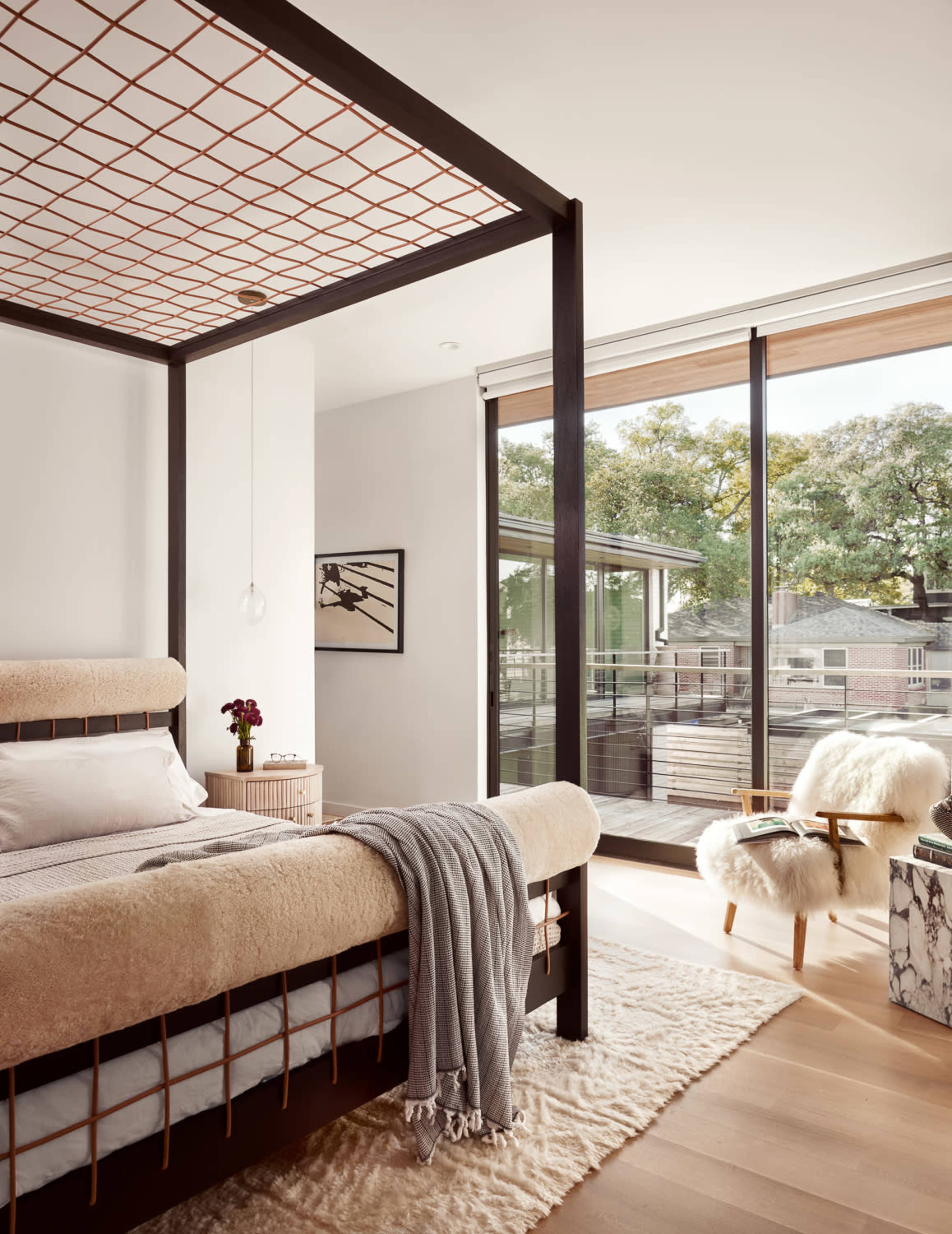 The bedroom features a modern four-poster bed with a light-colored bedspread, accompanied by a cozy throw, a plush rug, and large windows overlooking a balcony.