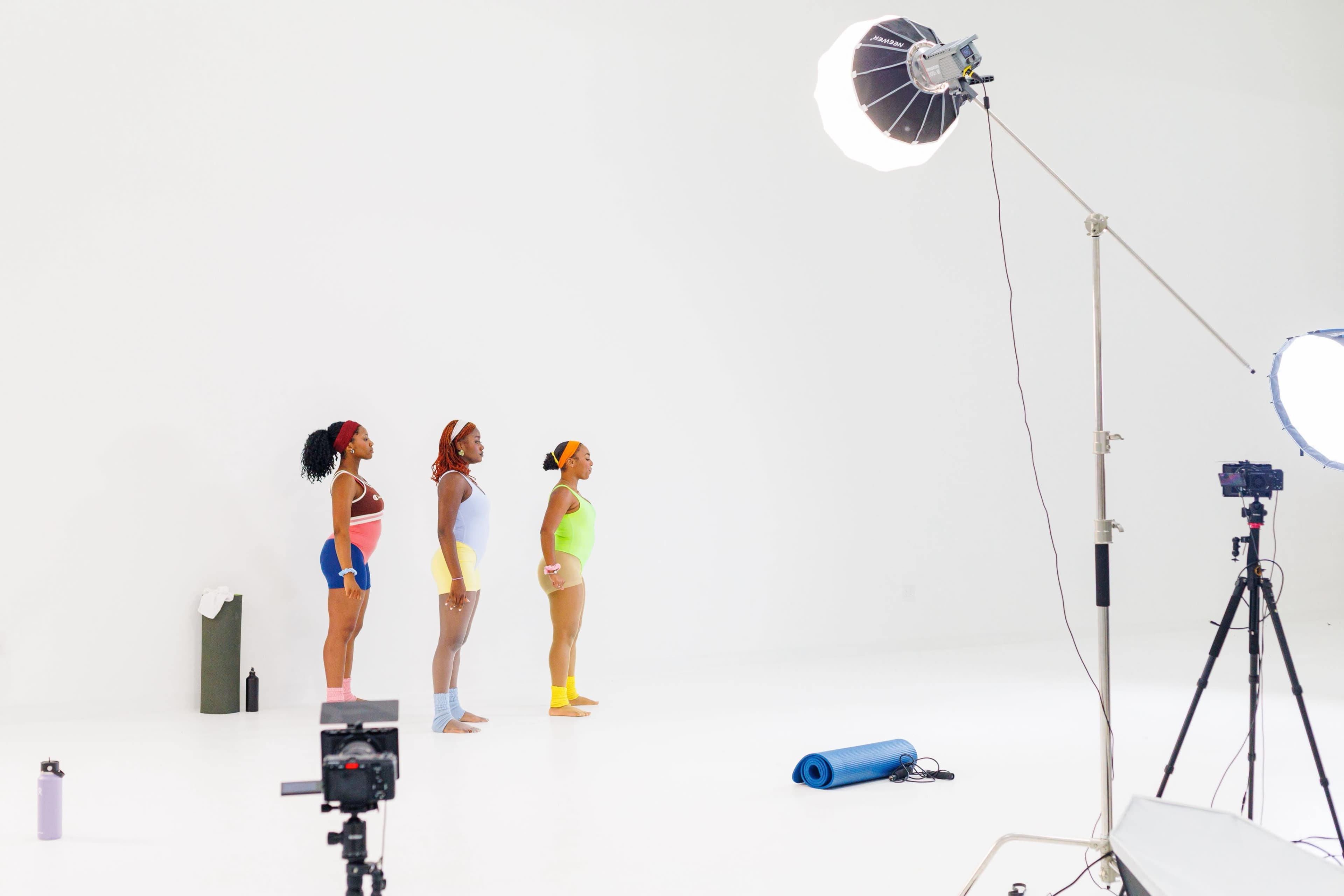 Three women in colorful athletic wear stand in a line while facing a camera in a brightly lit studio.