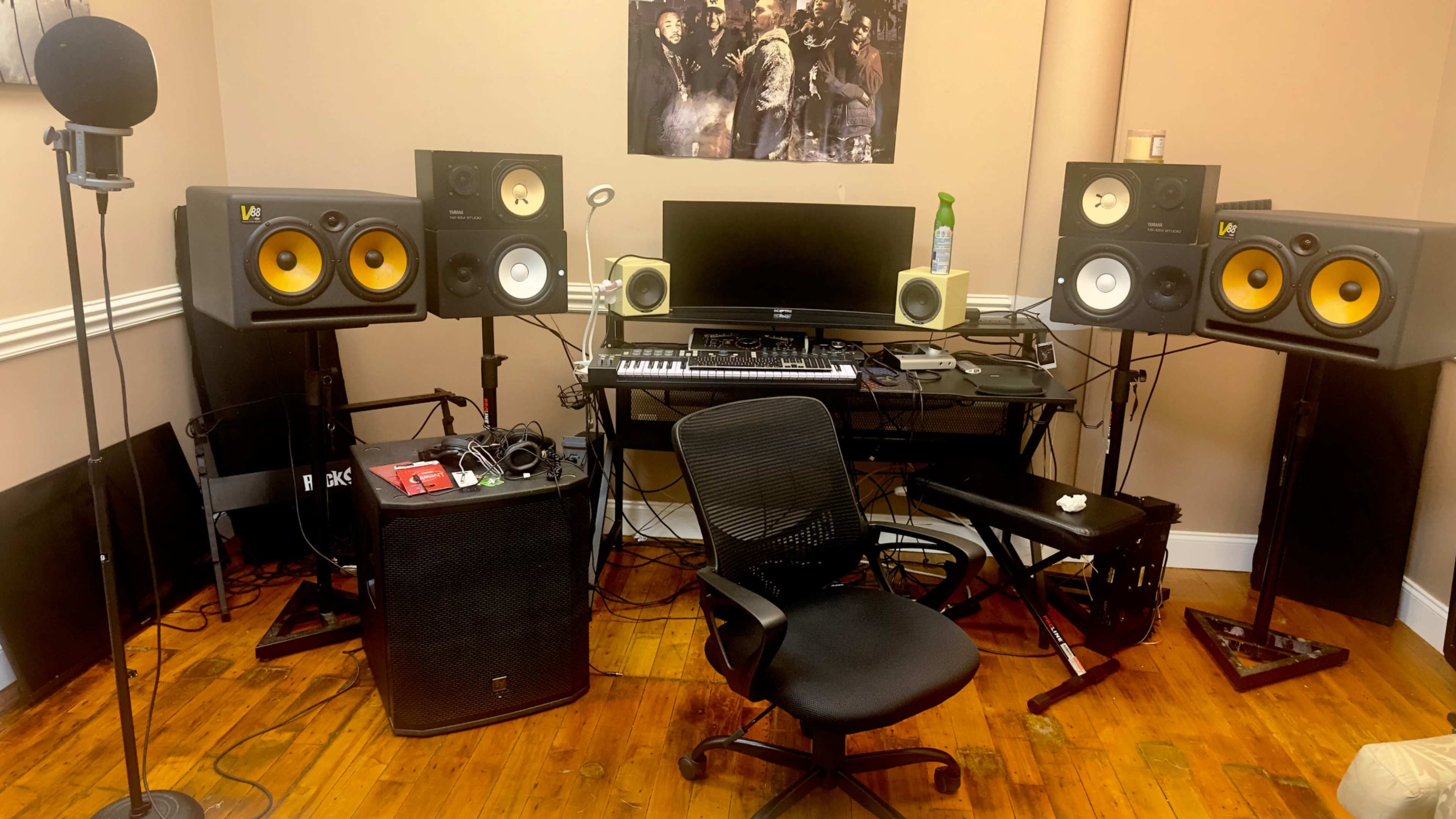 The image shows a music production studio with a microphone, speakers, a computer monitor, and various musical equipment arranged around a black chair.