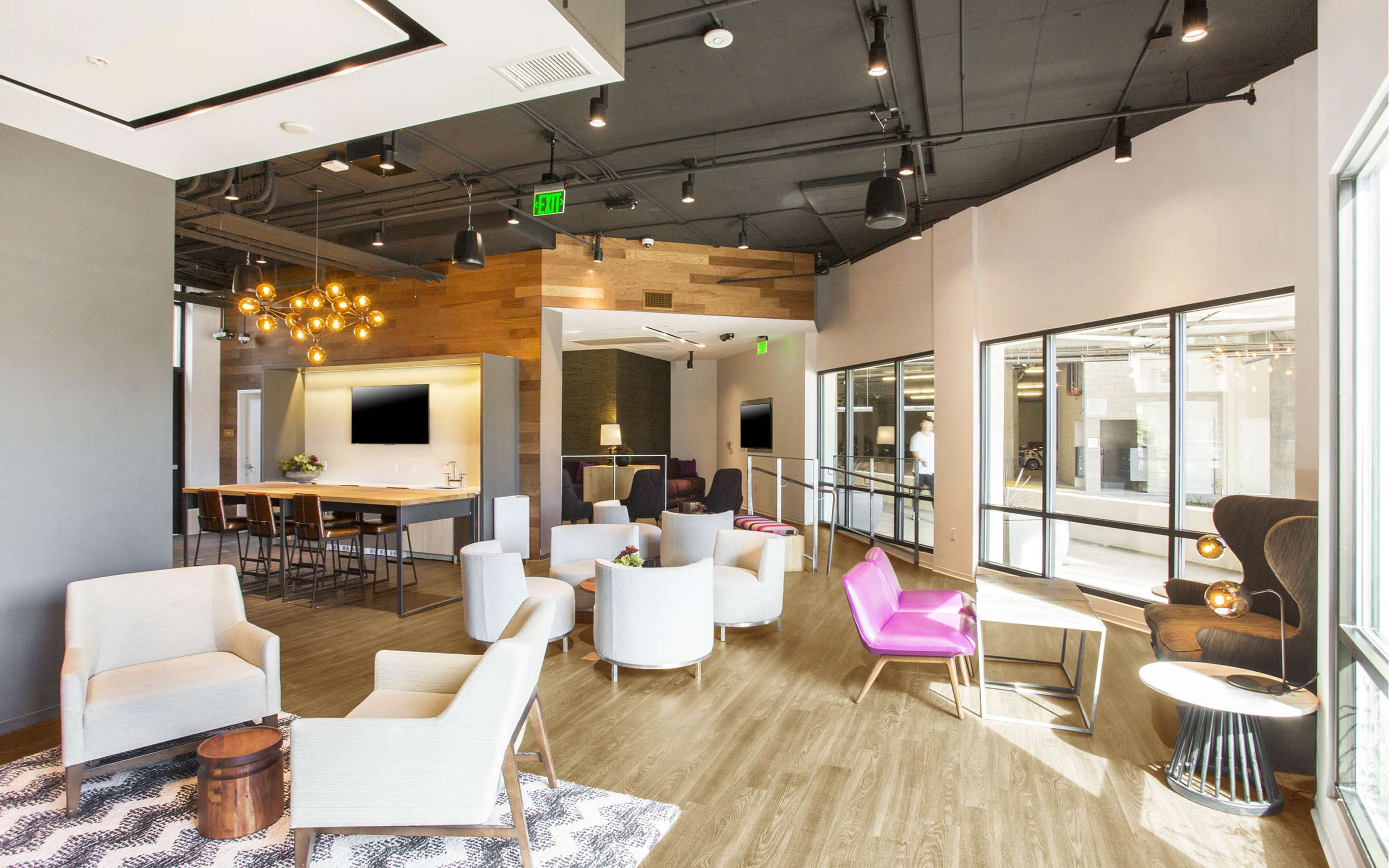 A modern lounge area with various seating options, including white armchairs and a pink chair, surrounded by large windows and wooden accents.