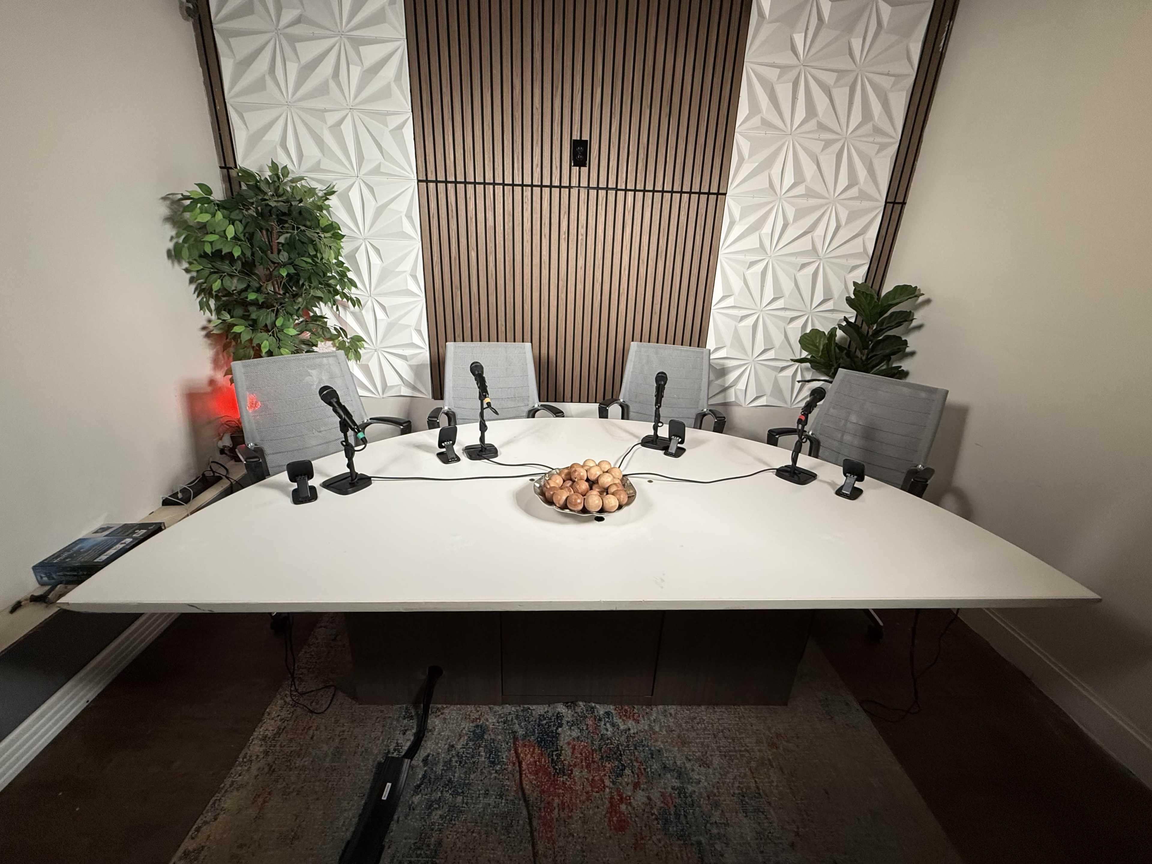 A round table with six microphones is set up in a small meeting room with decorative plants and a bowl of eggs in the center.