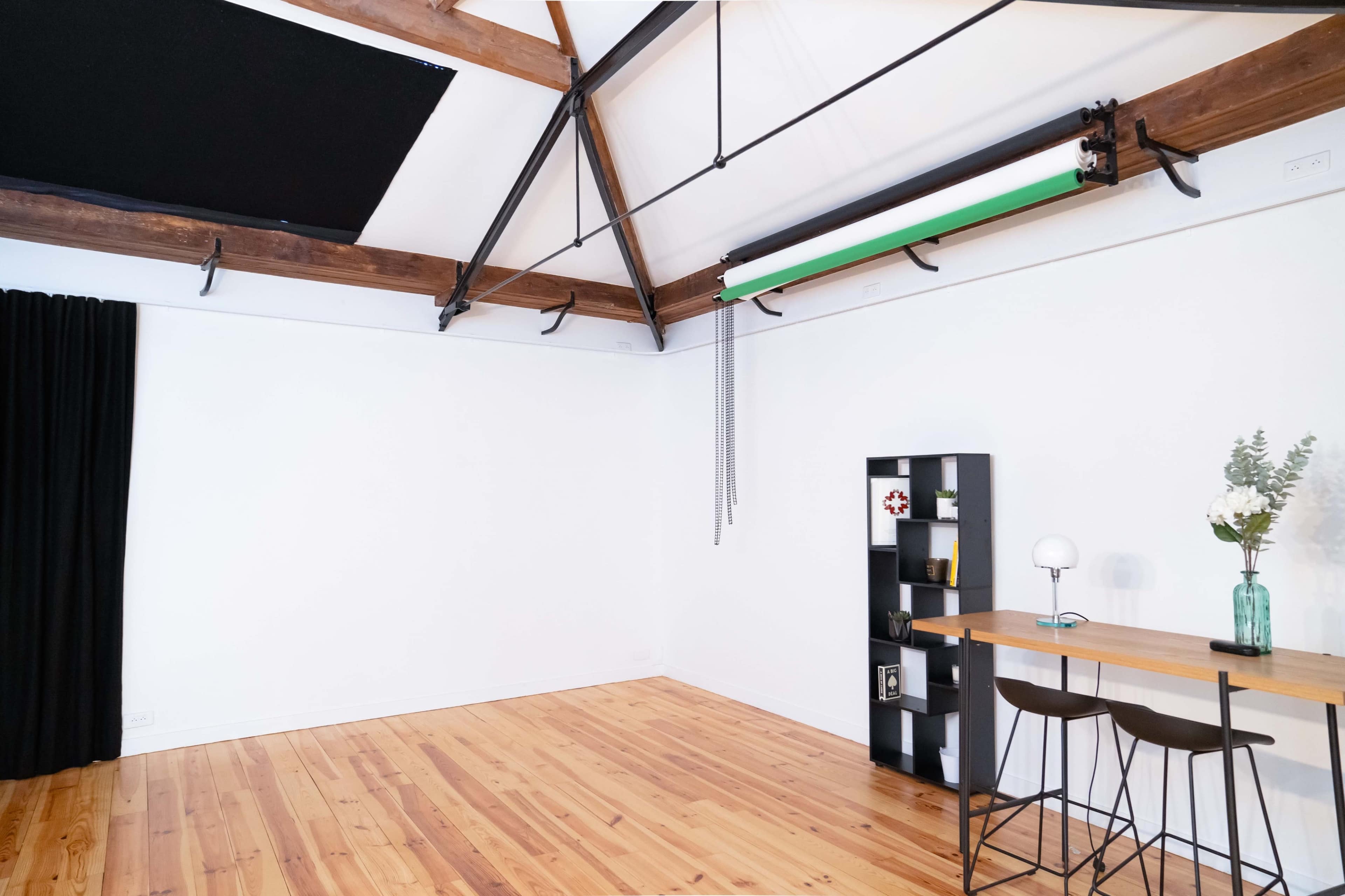 A bright, minimalistic room with wooden floors, a black shelving unit, a table with two chairs, and a large green backdrop hanging from the ceiling.
