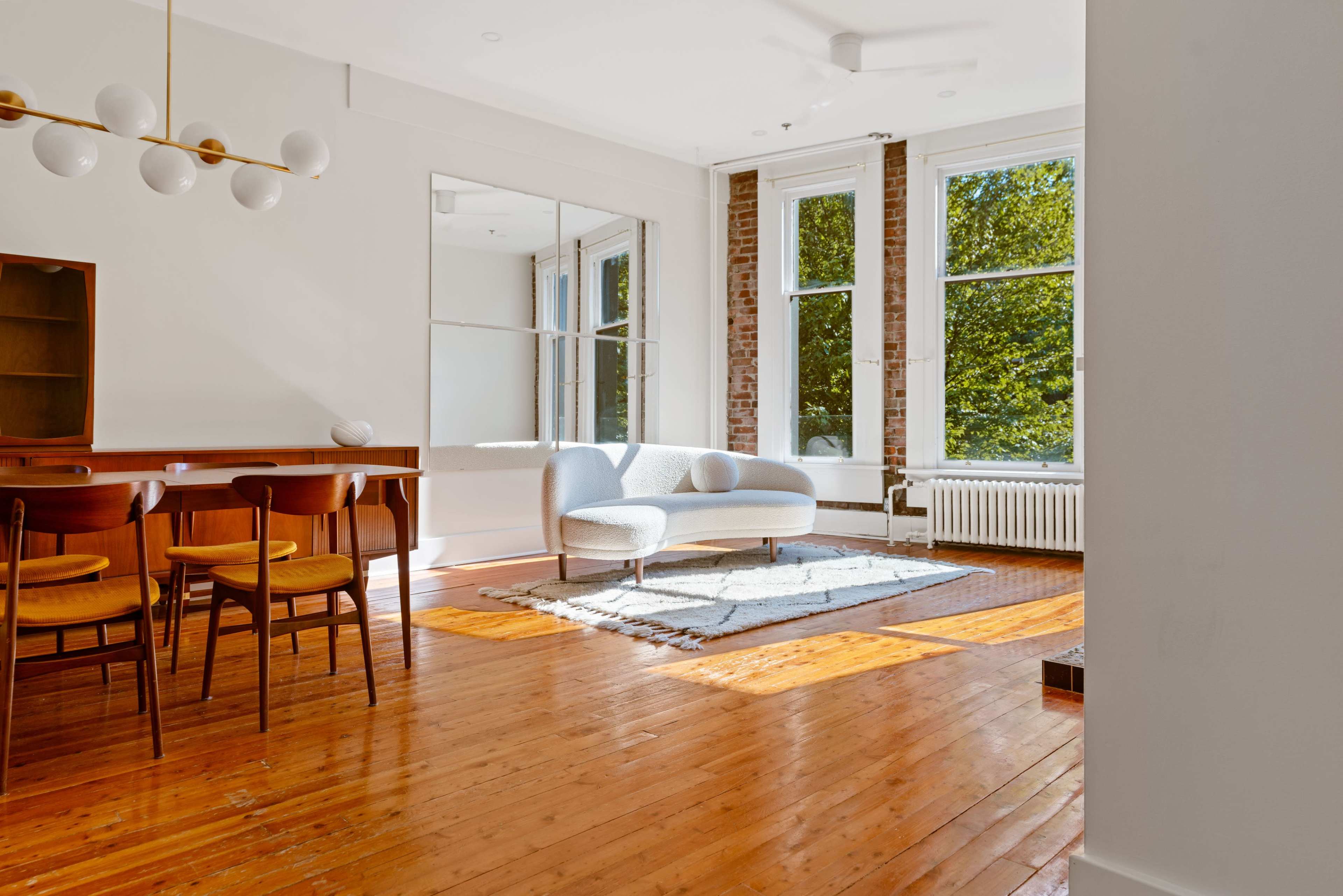 A bright living space with a dining table and chairs, a light-colored sofa, large windows, and exposed brick walls.