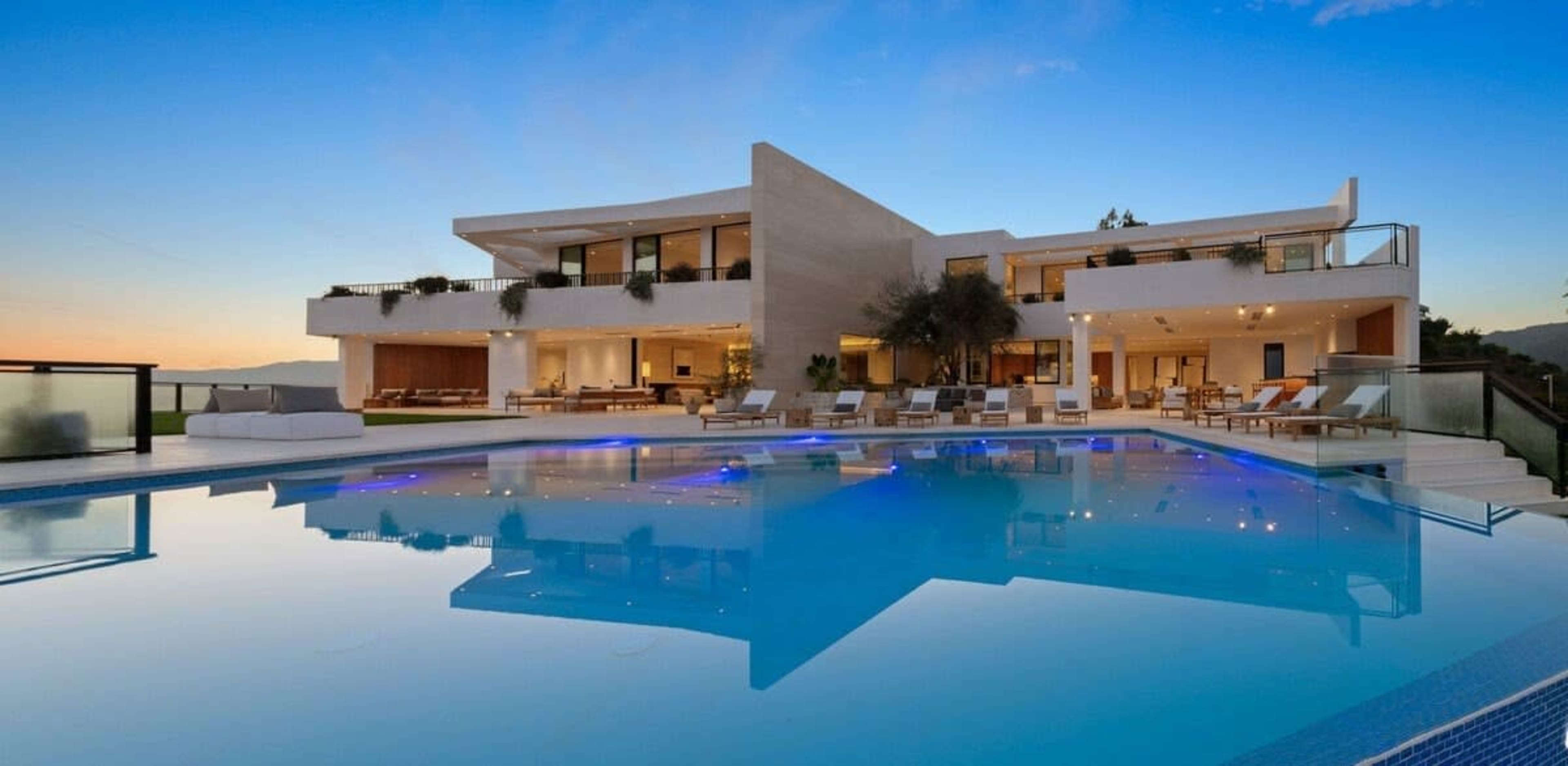 A modern, multi-level villa with a large swimming pool reflects the twilight sky and is surrounded by lounge chairs and greenery.
