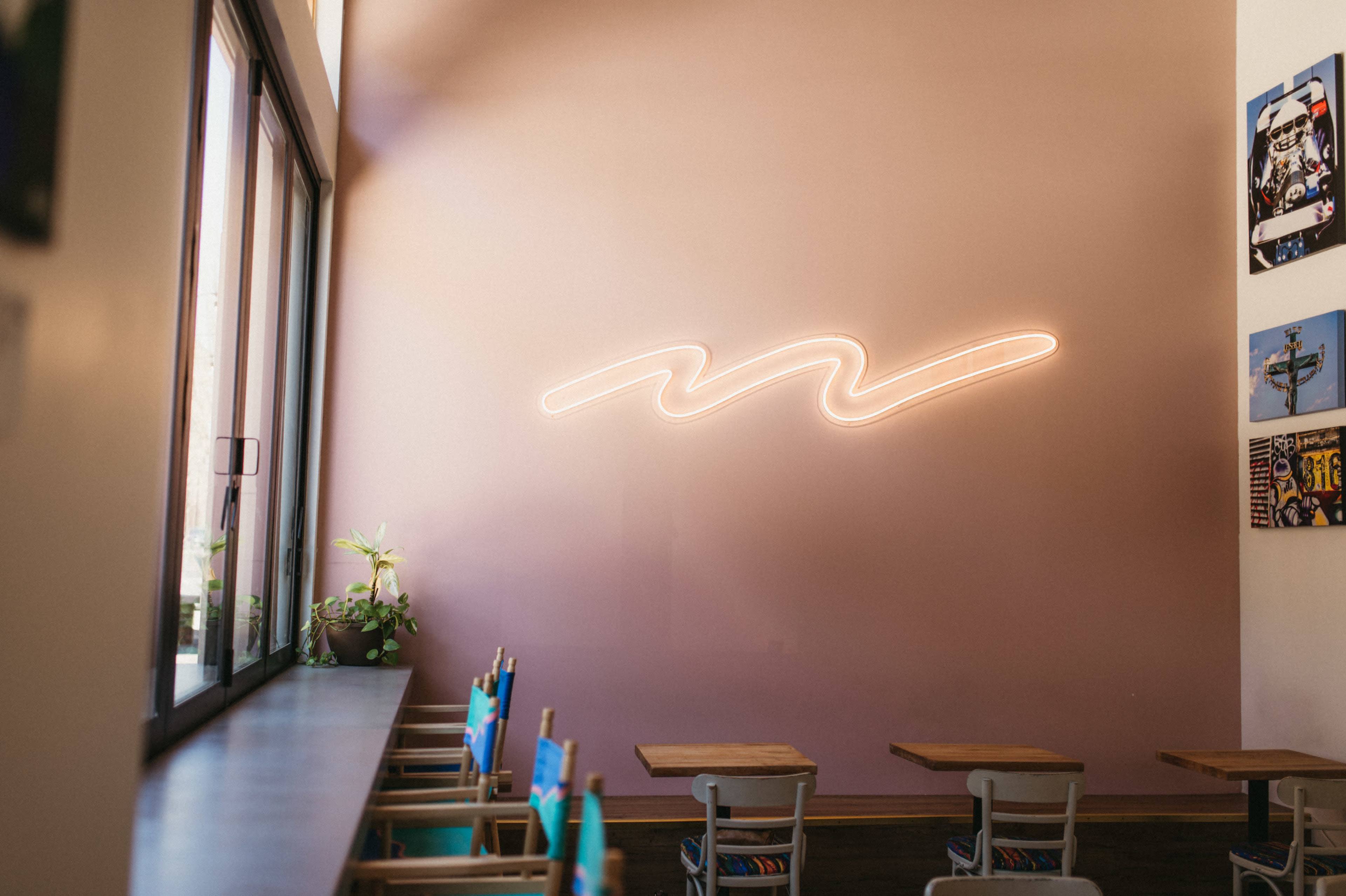 A café interior features a pink wall with a wavy neon light above a row of tables and chairs.
