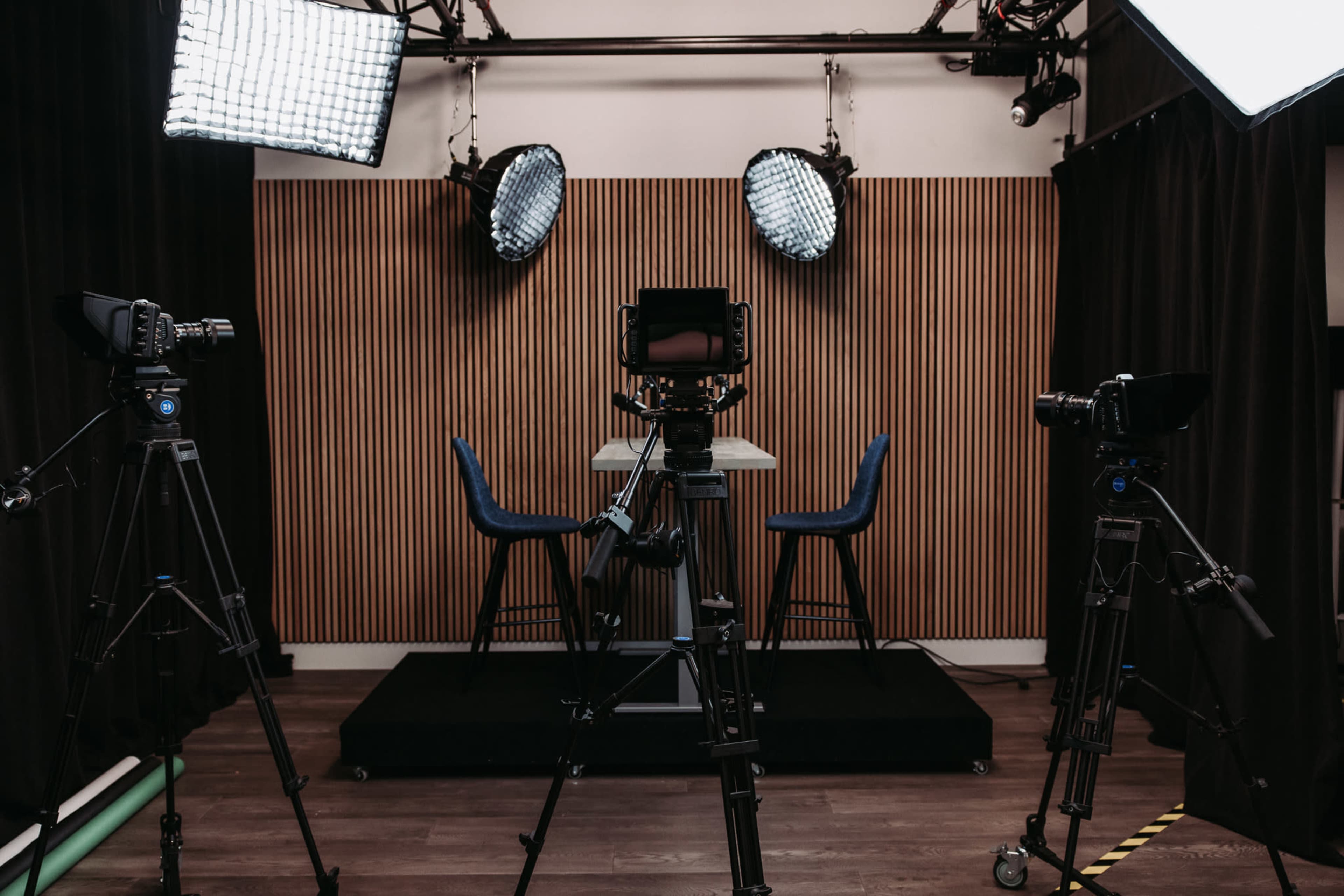 The image shows a video production studio setup with multiple cameras and lights directed at a table with two chairs against a wooden paneled background.
