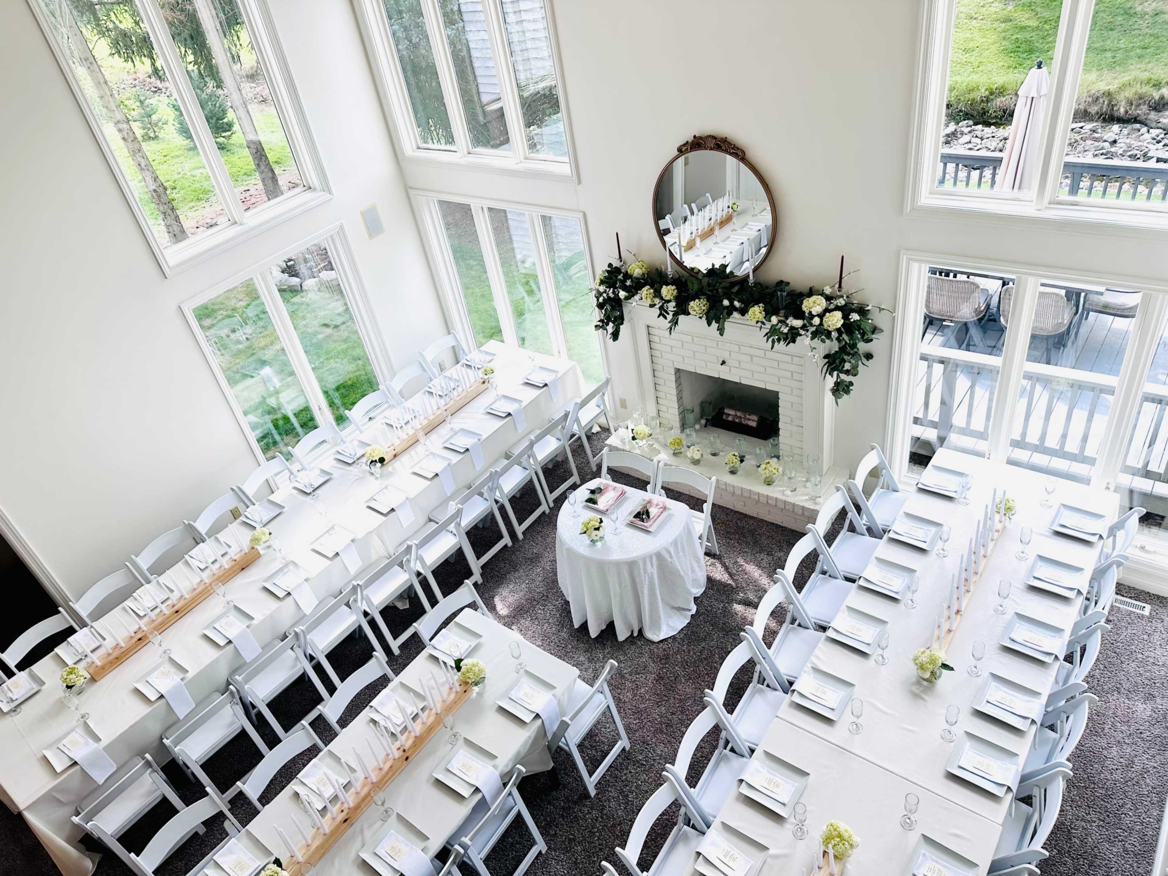 A spacious, elegantly arranged dining area features long tables set for an event, with a centerpiece table and large windows allowing natural light to illuminate the space.