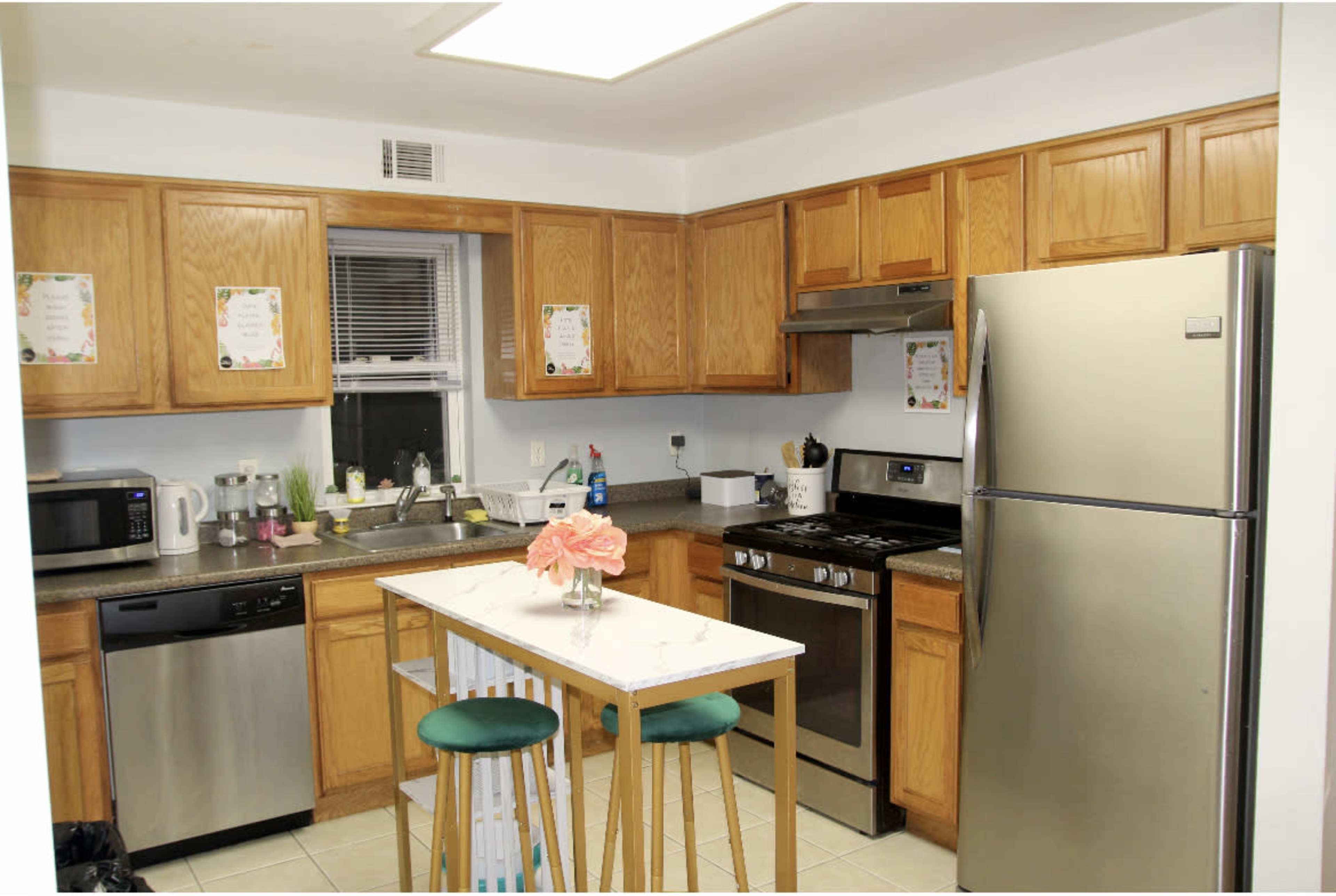 The image shows a kitchen with wooden cabinets, stainless steel appliances, and a small island with stools in the center.