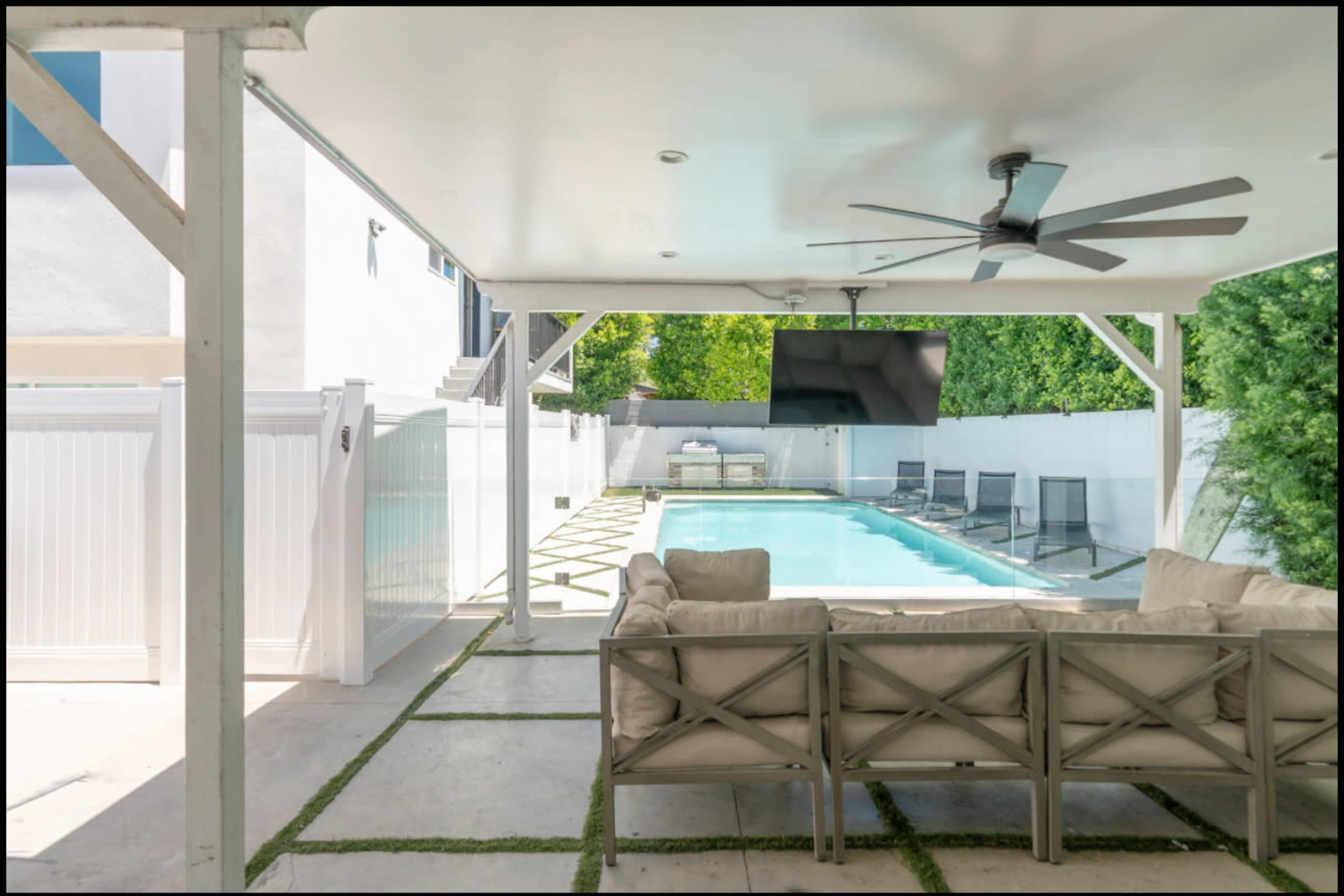 The image shows a covered outdoor patio area with a seating arrangement facing a swimming pool, flanked by greenery and outdoor furniture.