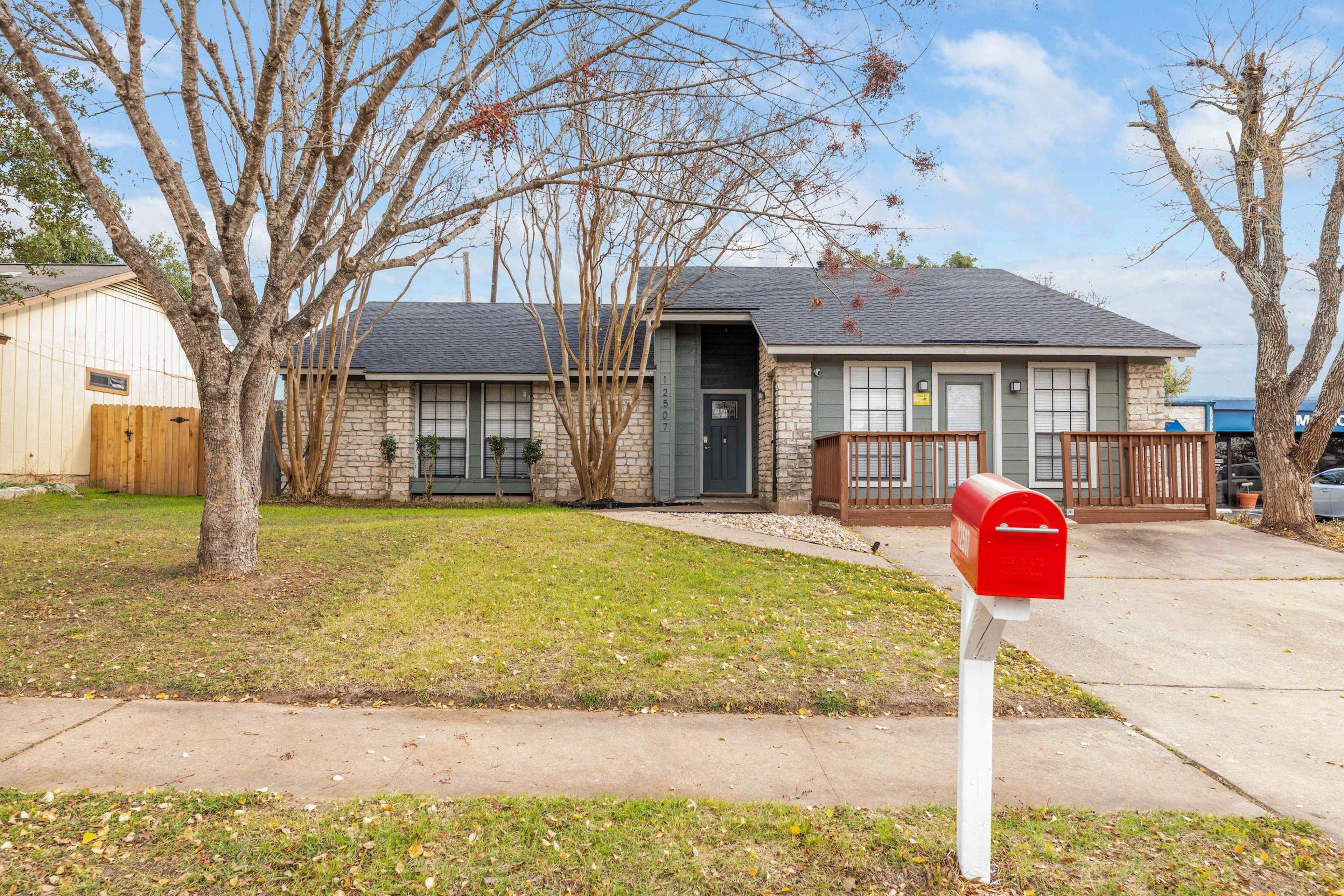 A single-story stone house with a dark roof sits on a grassy lot, featuring a wooden deck and a bright red mailbox at the edge of the driveway.