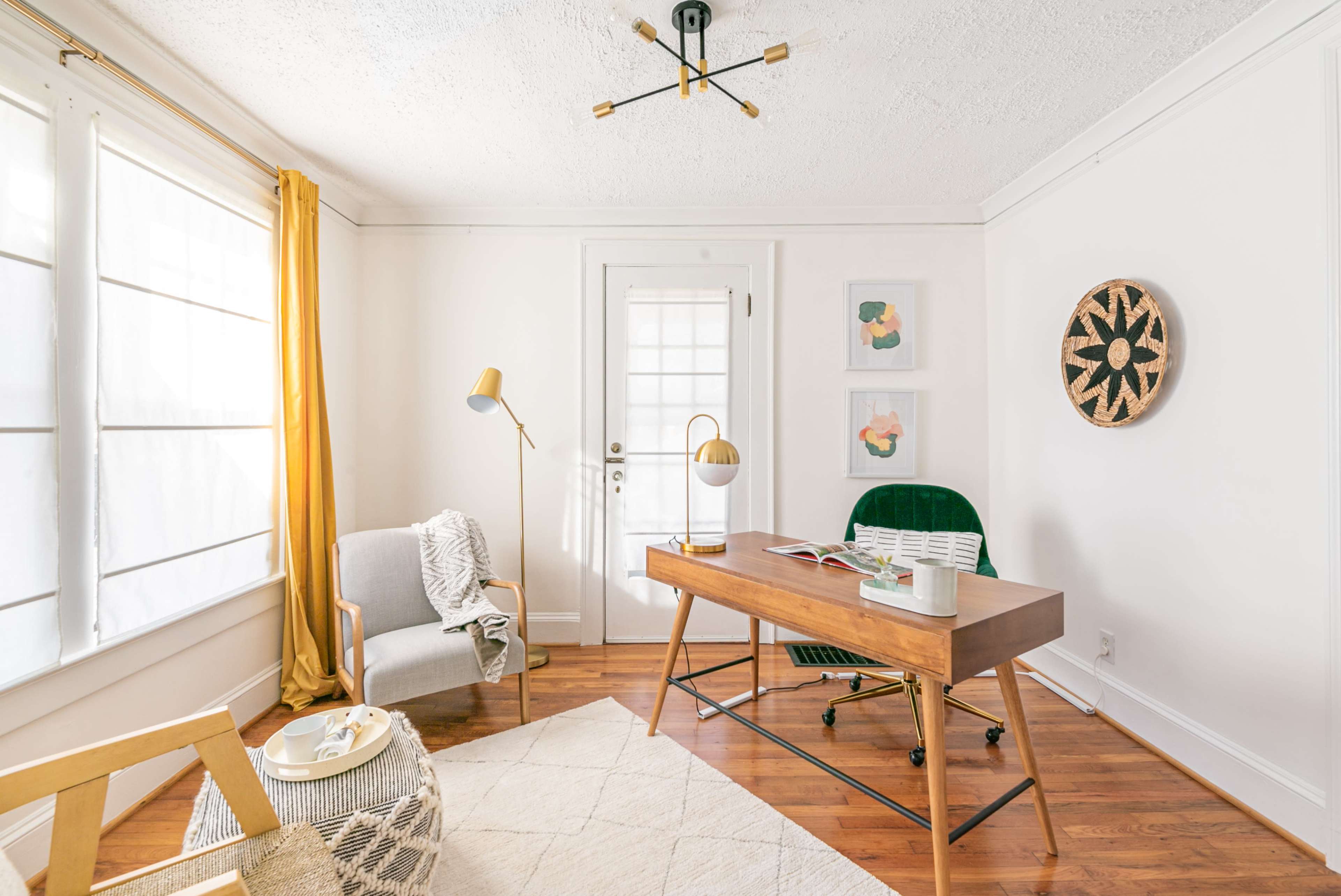 A modern office space features a wooden desk, a green chair, a cozy reading nook with a gray armchair, and large windows with yellow curtains.