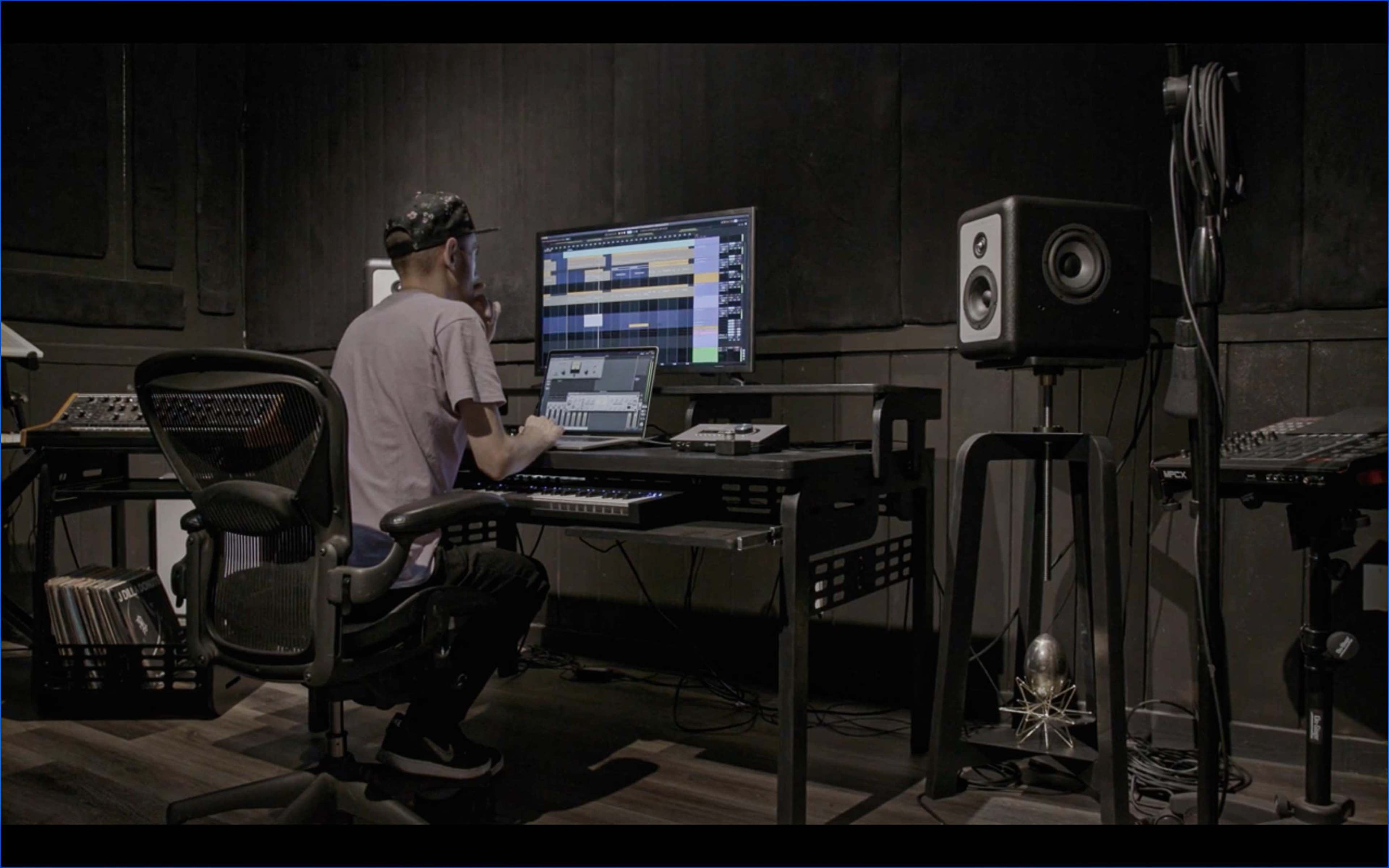 A person sits at a desk in a music studio, focusing on a computer screen and surrounded by audio equipment.