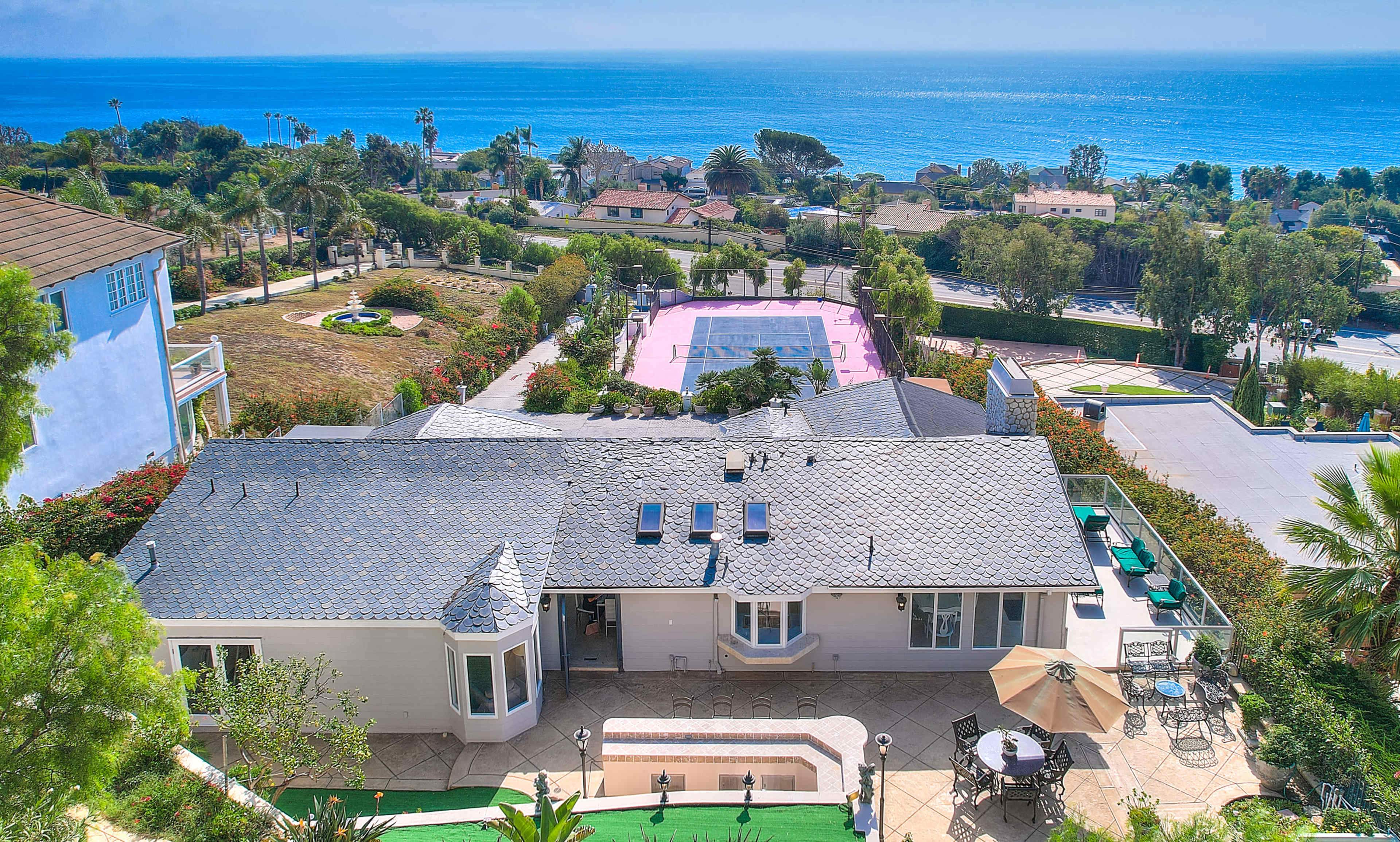 A spacious house overlooking the ocean, with a tennis court and lush landscaping in the background.