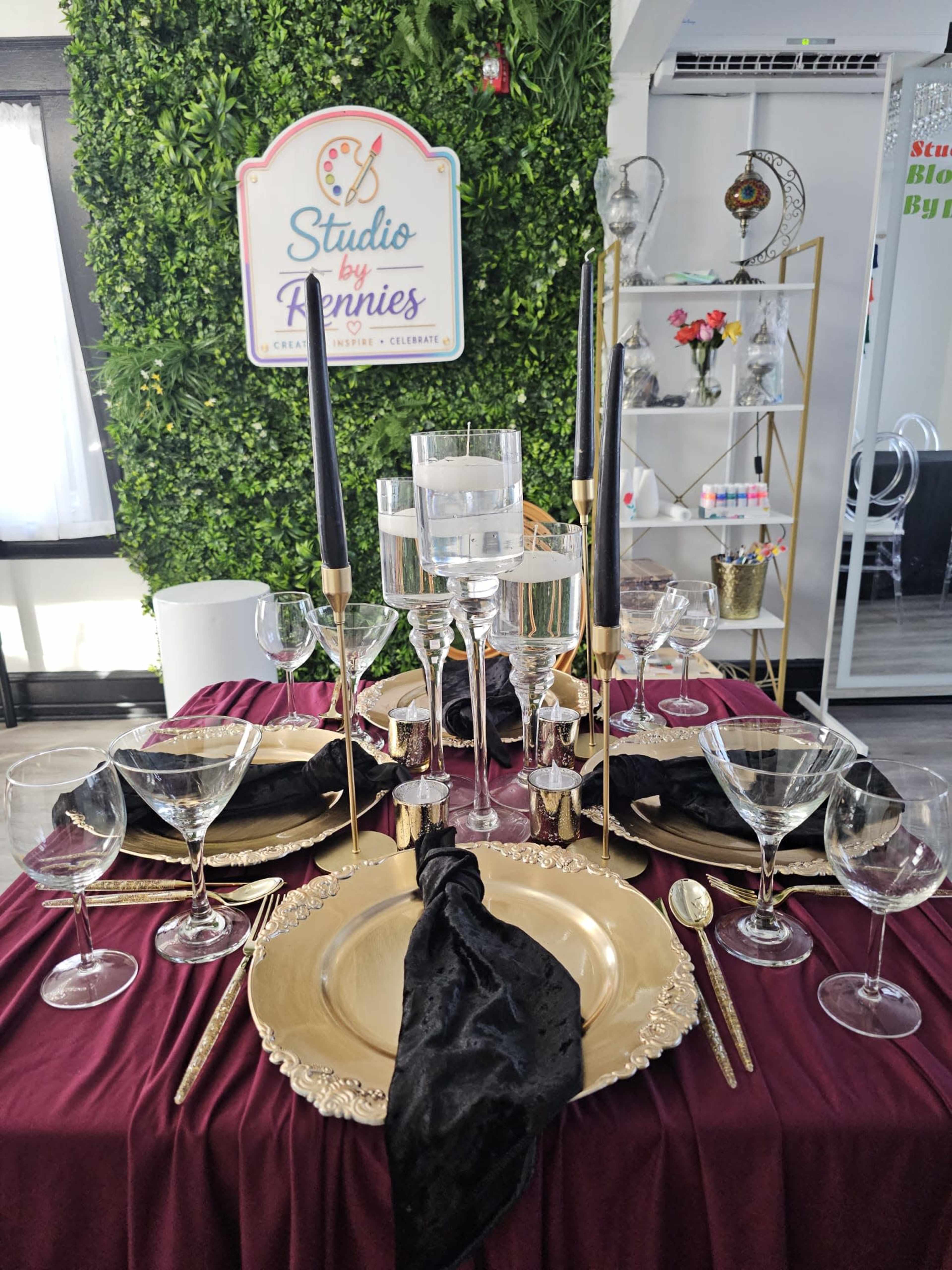 A formal dining table is set with elegant tableware, including gold plates, crystal glassware, and tall black candles, against a backdrop of a greenery wall and a decorative sign.