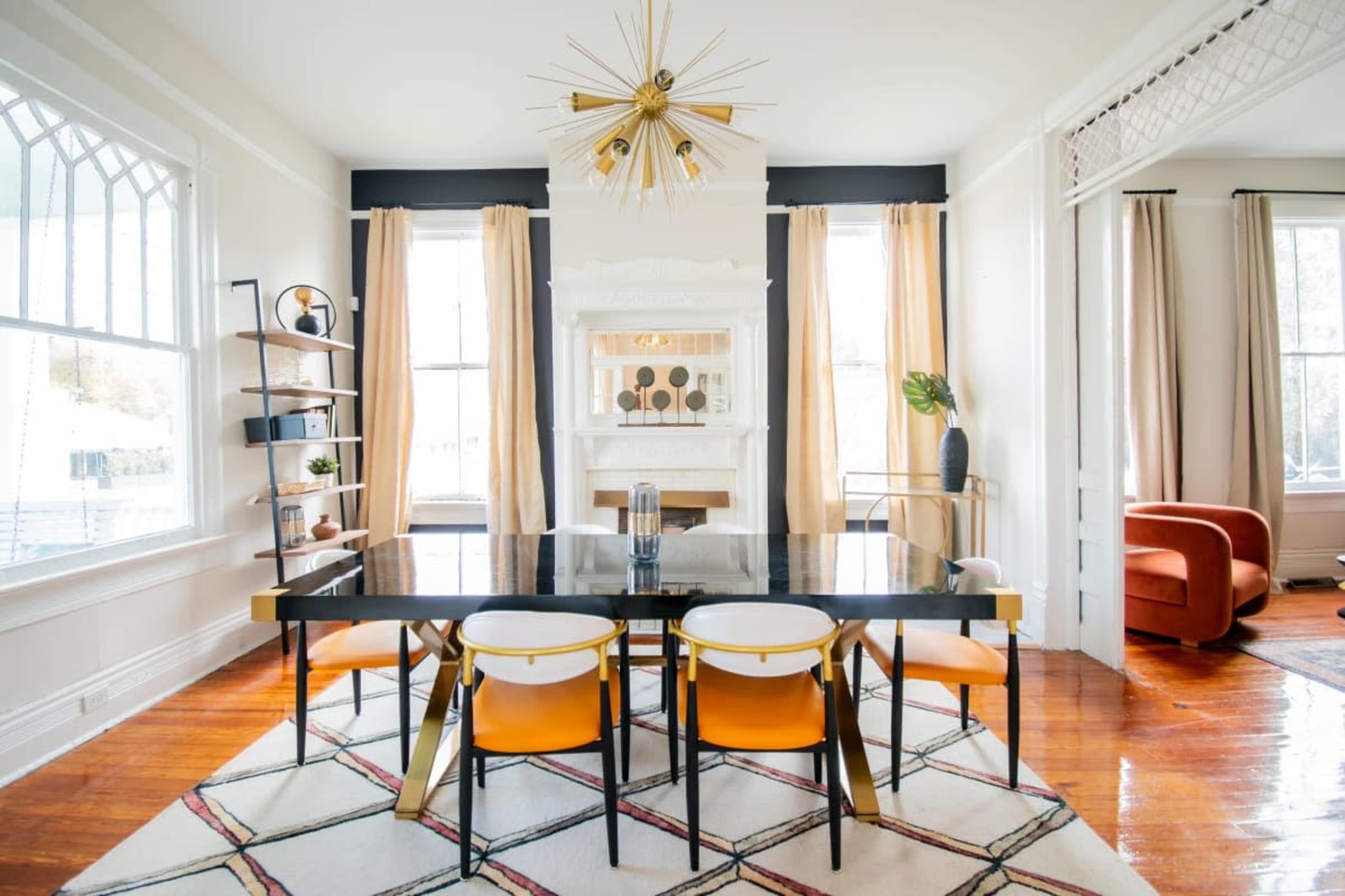 A modern dining room featuring a black dining table with orange-accented chairs, large windows with light curtains, and a fireplace surrounded by decorative shelves.
