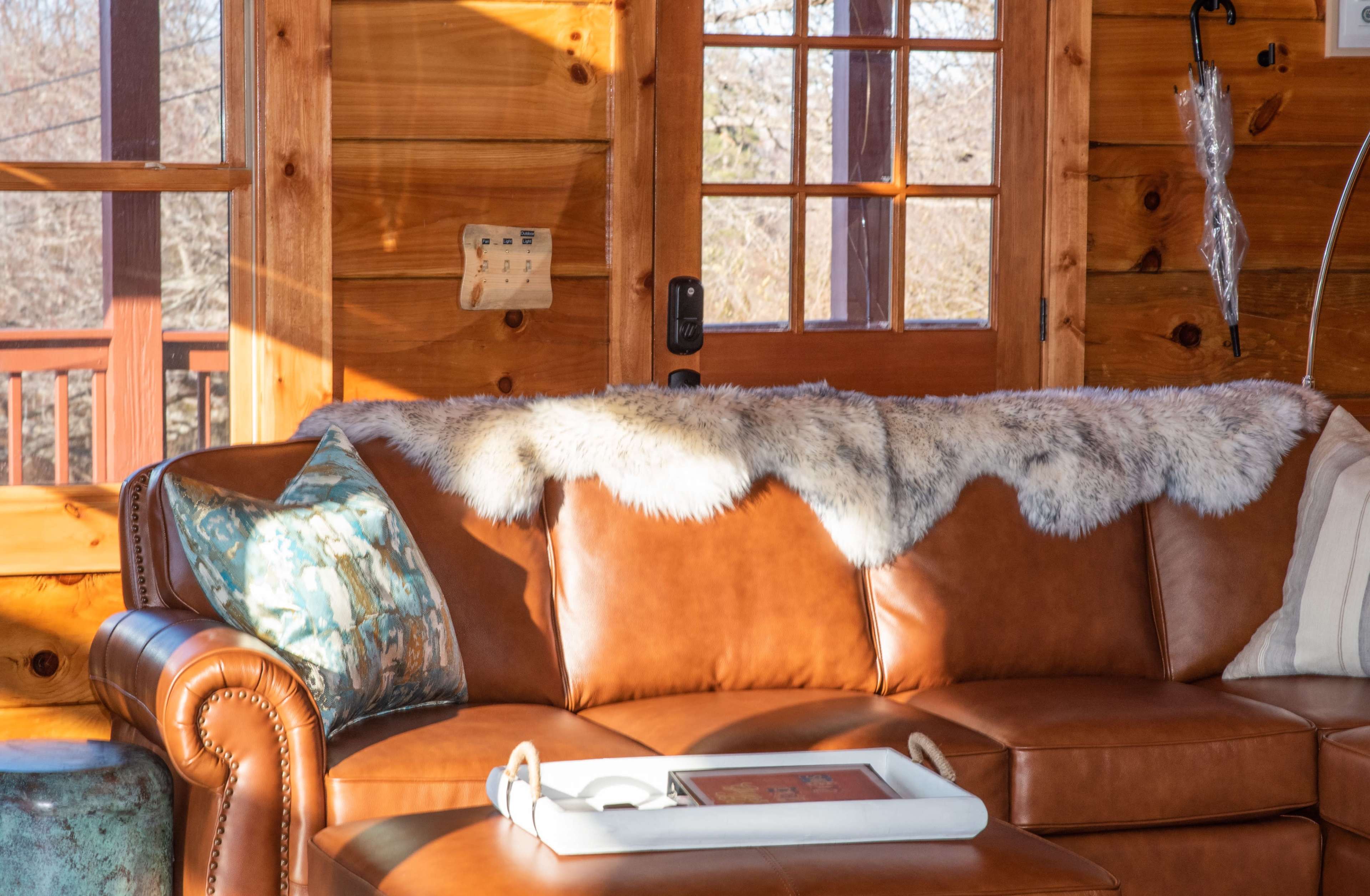 A leather couch is positioned in a wooden cabin, adorned with a faux fur throw and decorative pillows, with a tray resting on its arm.