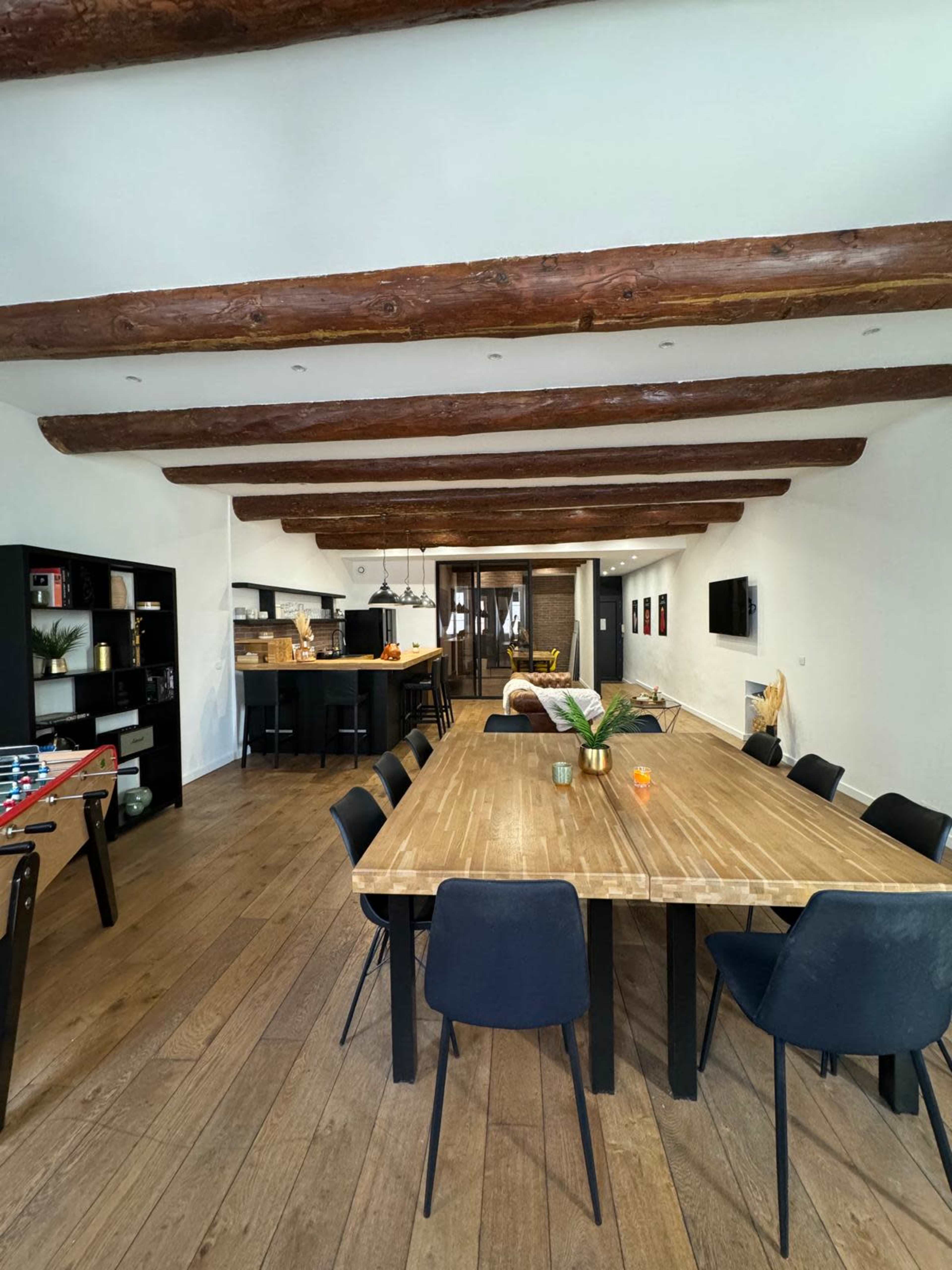 A spacious dining area features a large wooden table surrounded by black chairs, with exposed wooden beams above and an open kitchen in the background.