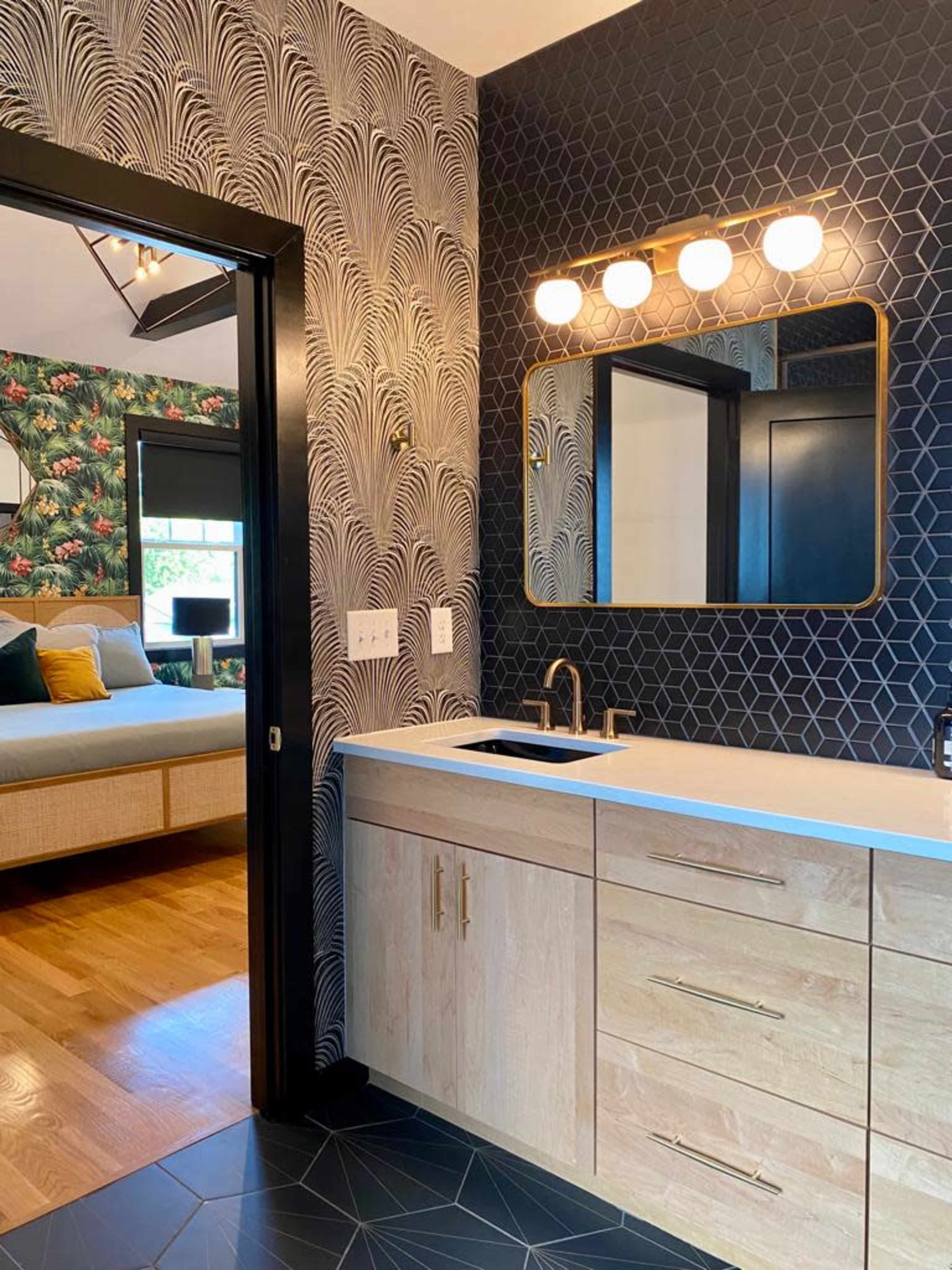 A modern bathroom featuring a vanity with a white countertop, wooden cabinets, a round mirror, and a geometric tile floor, adjacent to a bedroom with floral wallpaper.