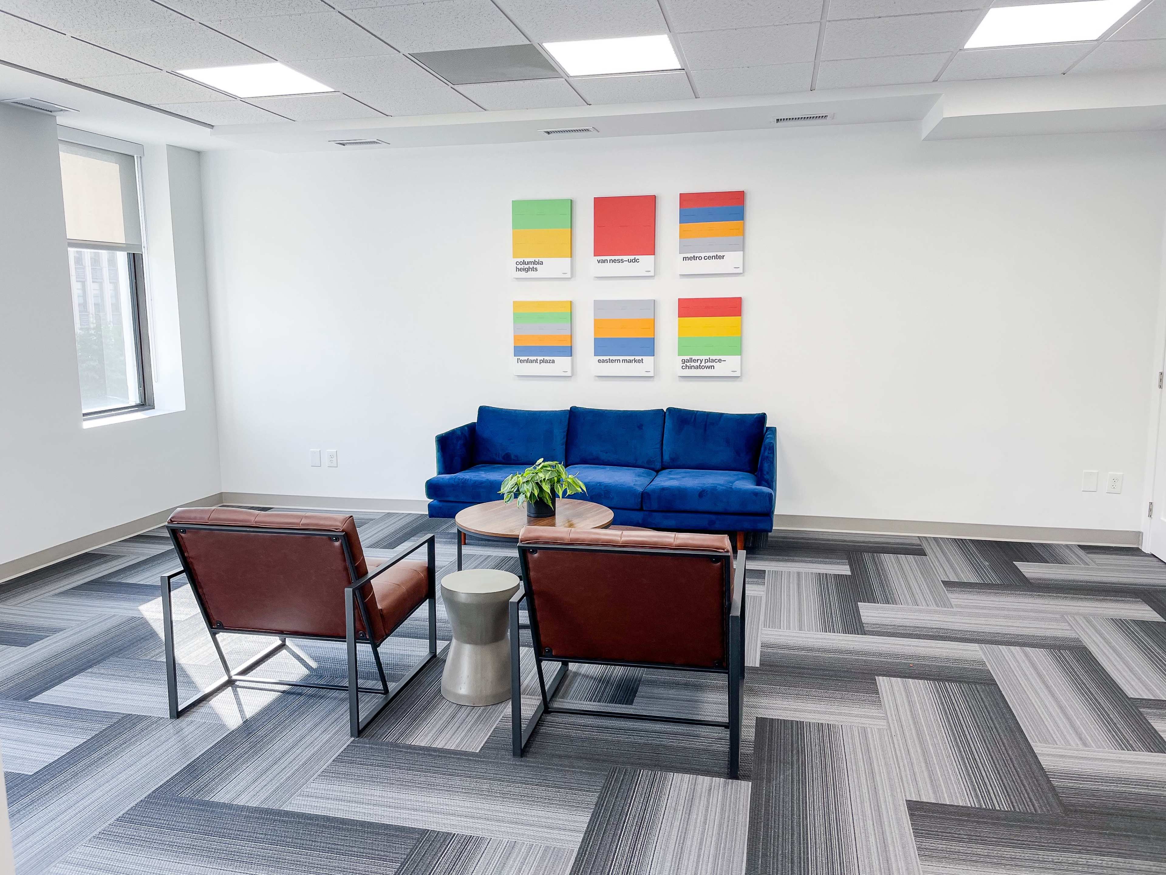 A modern office lounge features a blue sofa, two brown chairs, a round table, and colorful artwork on the wall.