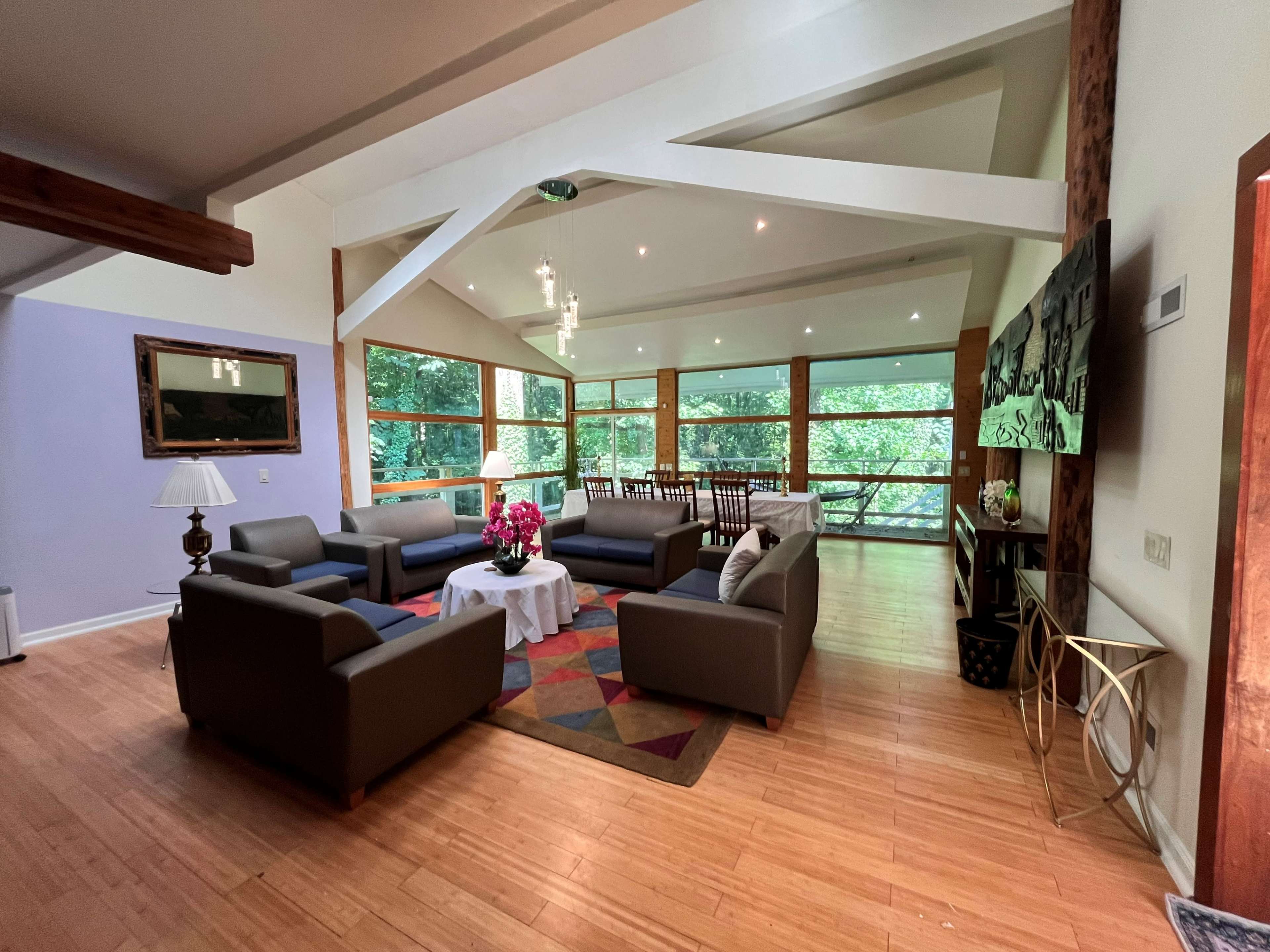 A spacious living area with large windows overlooking greenery, featuring modern sofas arranged around a coffee table and a dining area in the background.