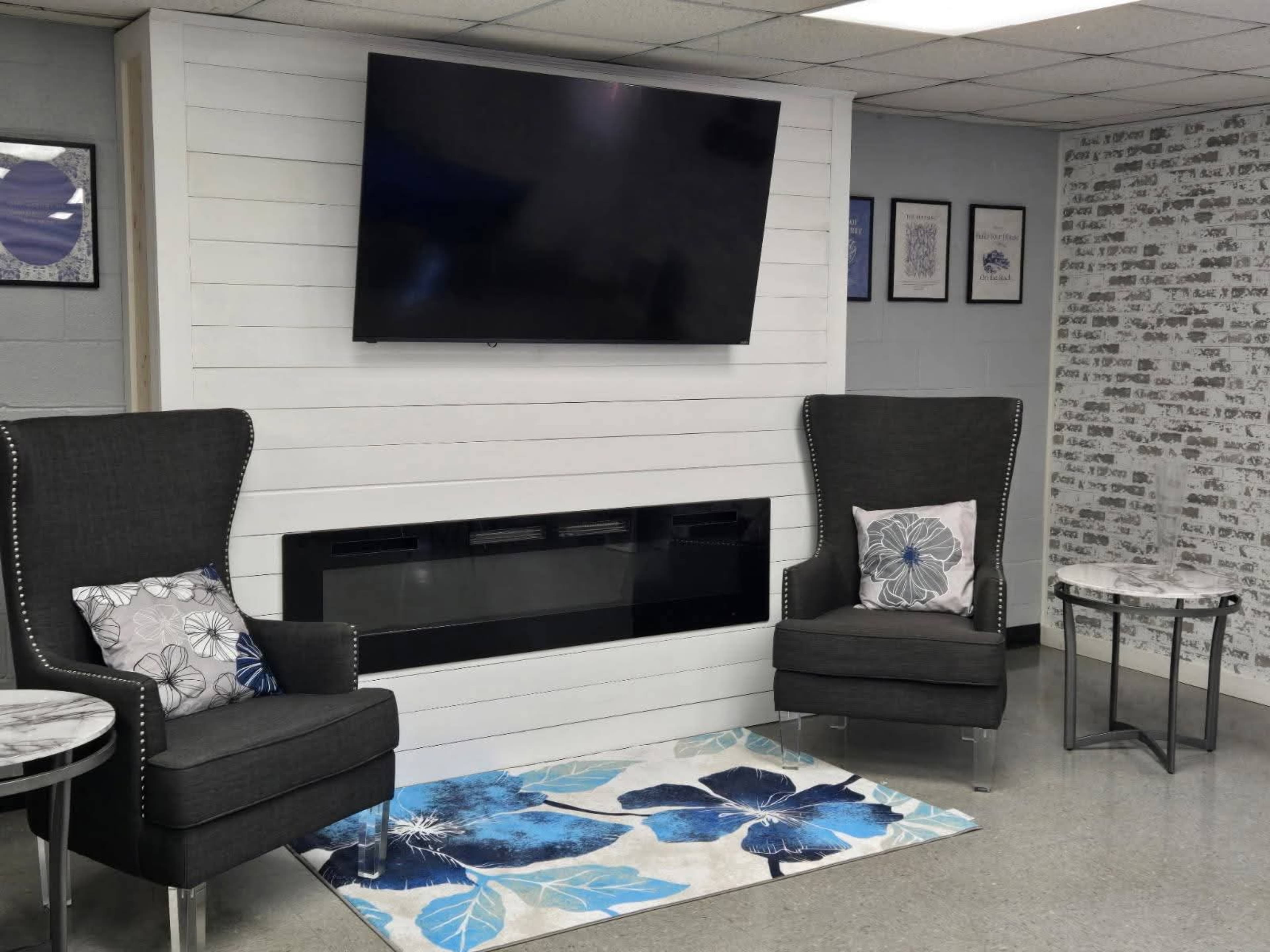 The image shows a modern seating area with two upholstered chairs flanking a sleek electric fireplace, a large wall-mounted television above, and a decorative rug featuring blue floral patterns on a light background.