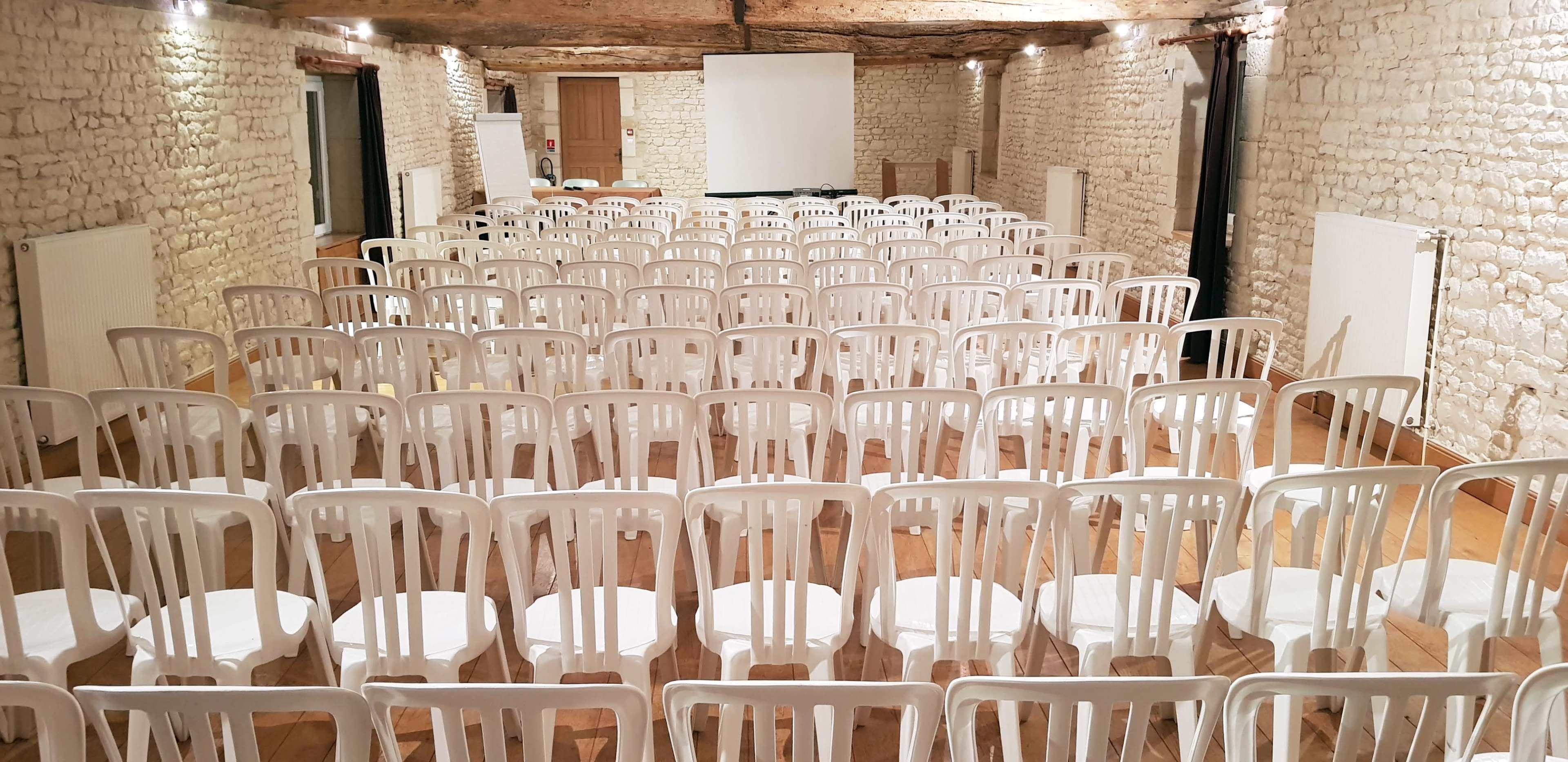 A room with stone walls and wooden beams contains rows of white plastic chairs arranged for an event.