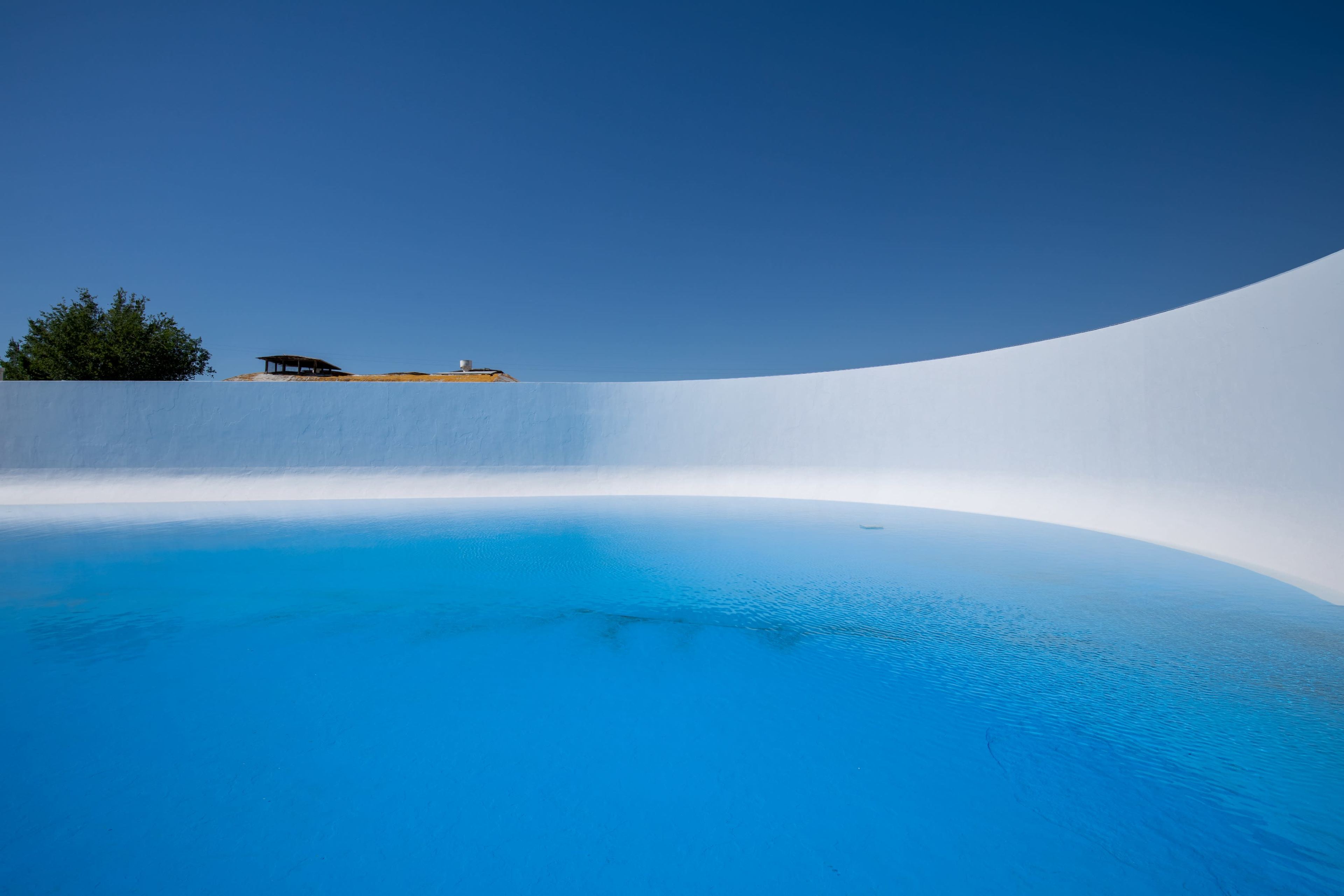 A curved white wall surrounds a tranquil blue swimming pool under a clear blue sky.