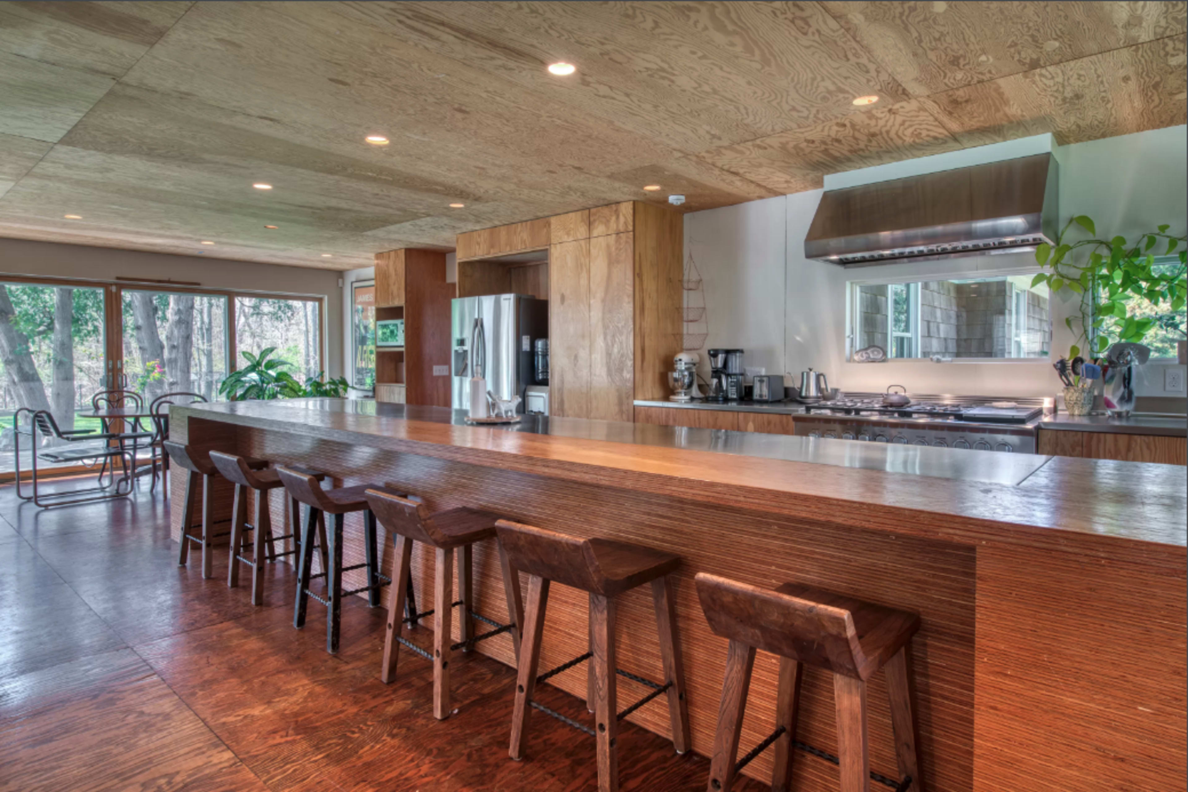 A modern kitchen featuring a long wooden bar with stools, a stove, and large windows overlooking a wooded area.