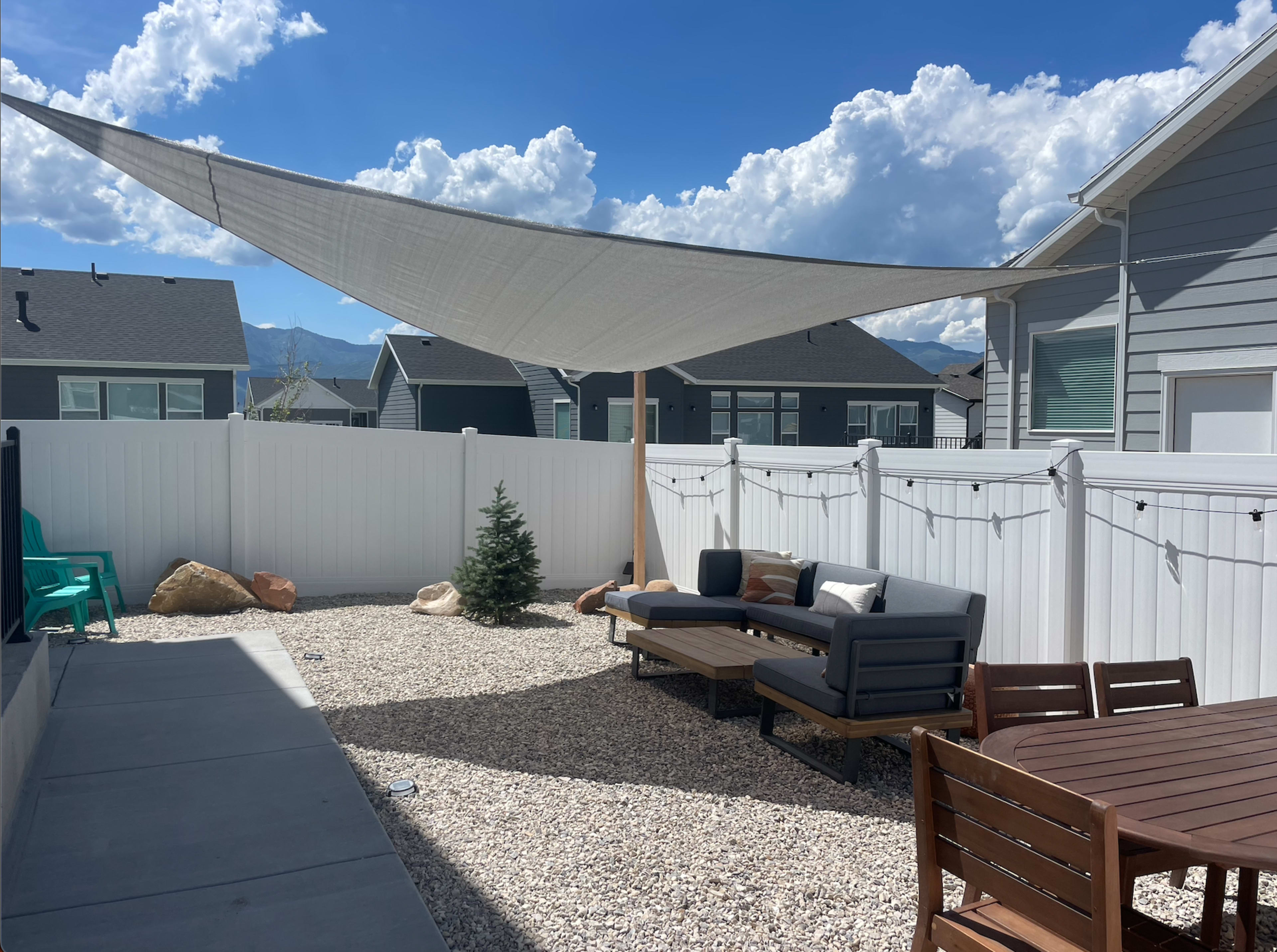 A gravel backyard with a shaded seating area, a small tree, and string lights, bordered by a white fence and houses in the background.