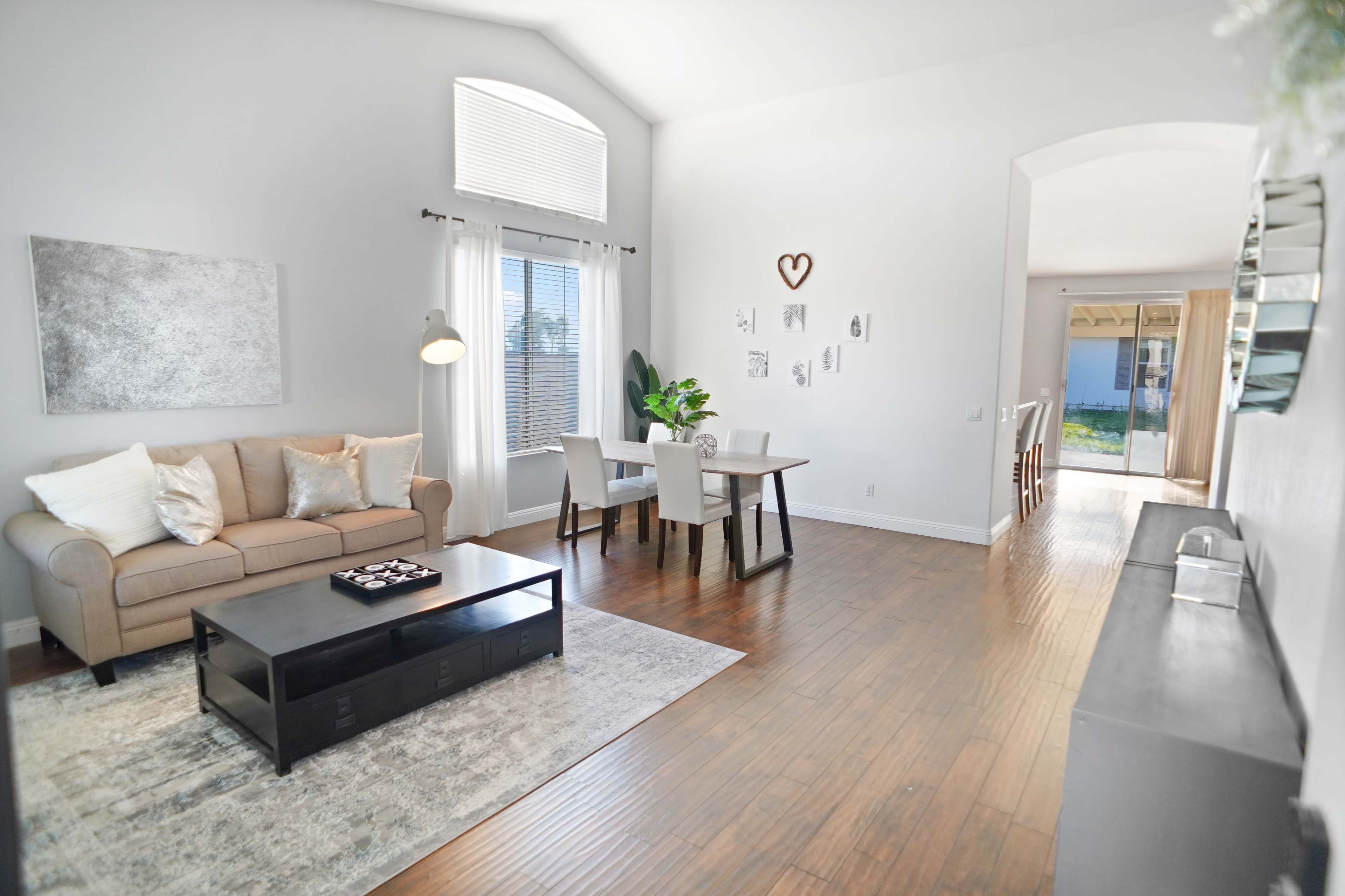 The image shows a spacious living area with a beige sofa, a black coffee table, a dining table set for four, and large windows allowing natural light.