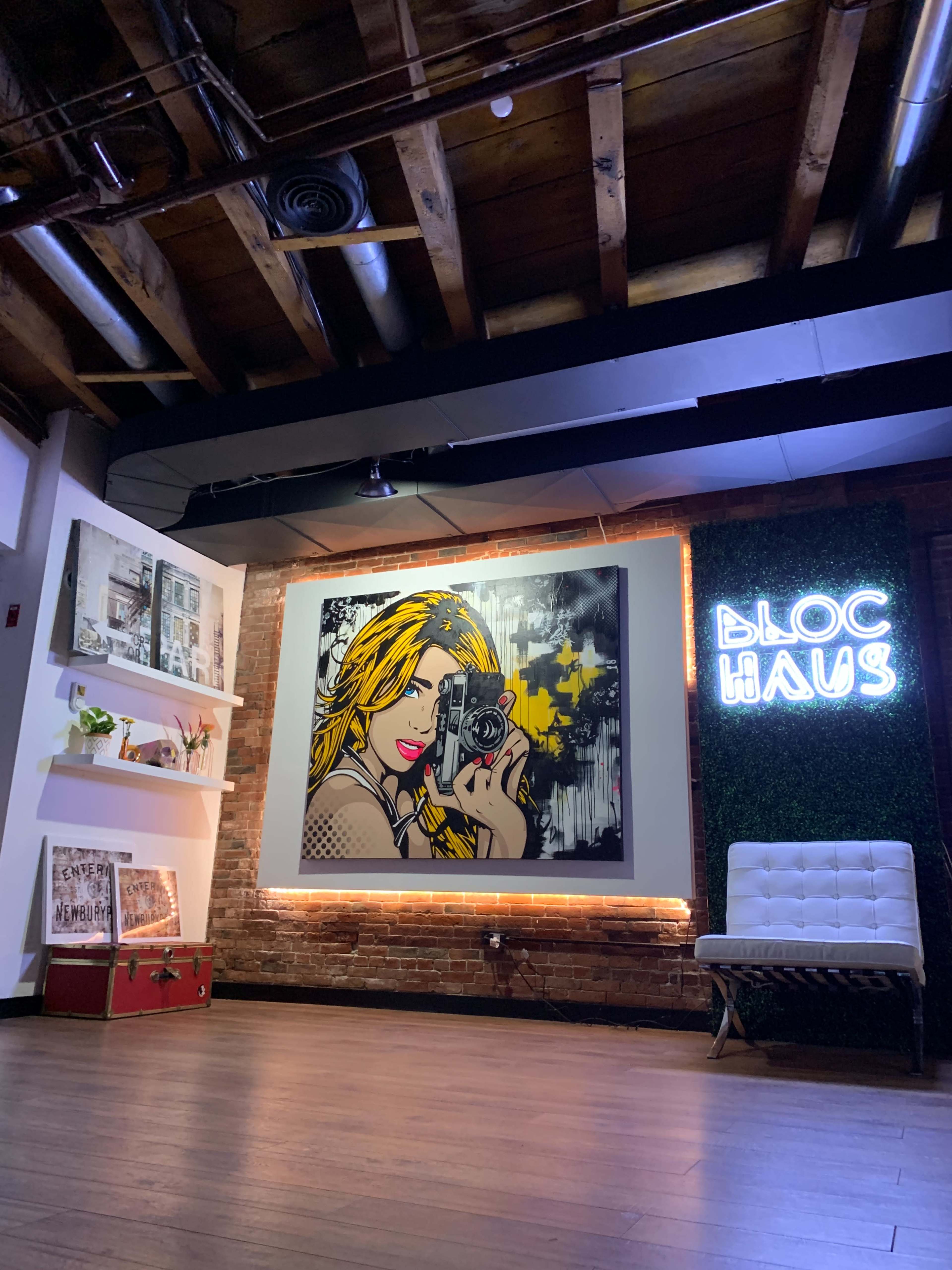 A vibrant mural of a woman with a camera on a wall in a stylish interior, alongside decorative shelves and a illuminated sign reading "BLOC HAUS."