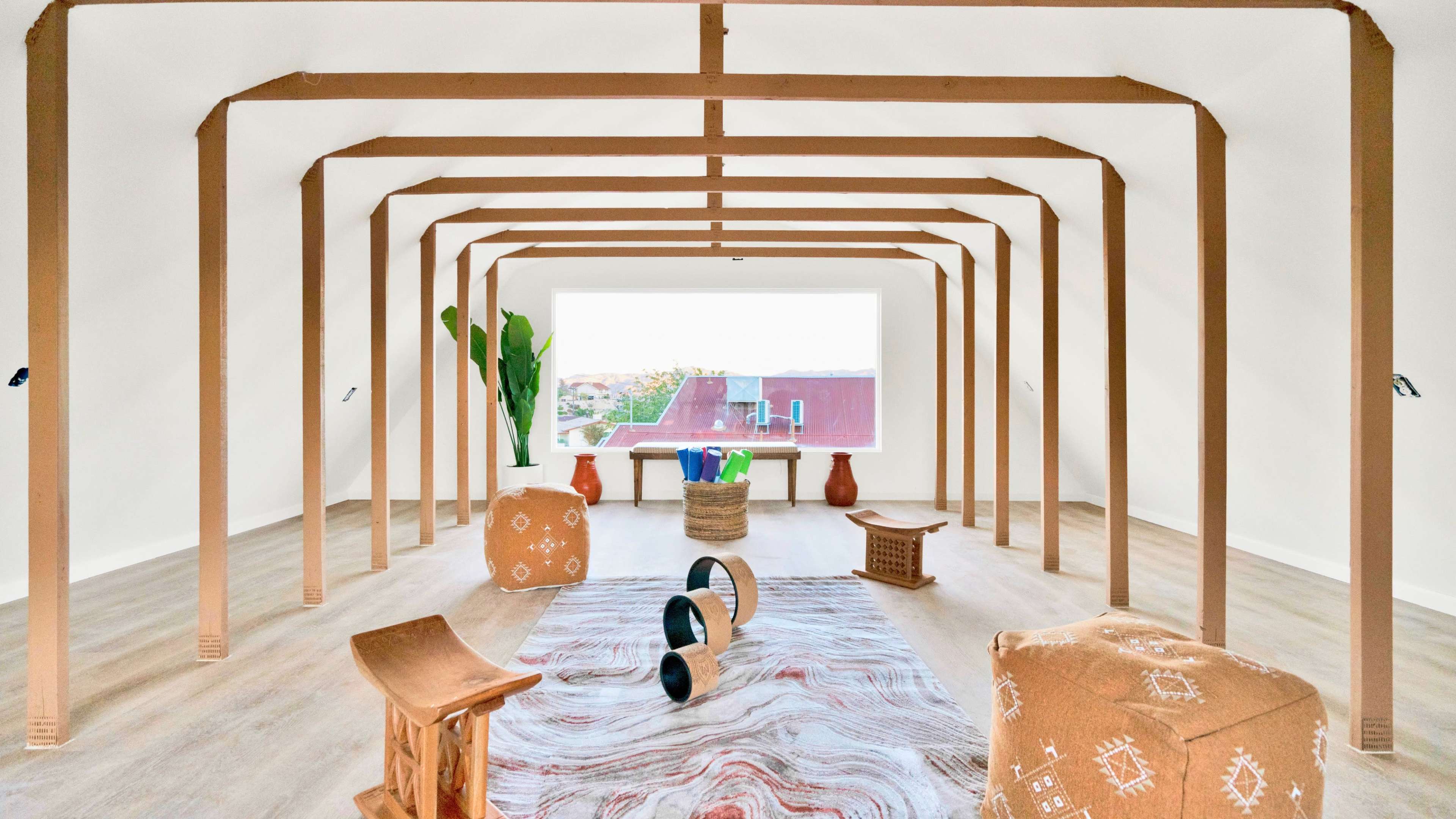 A spacious, minimalistic room with wooden beams, various furniture pieces, a large window, and plants.