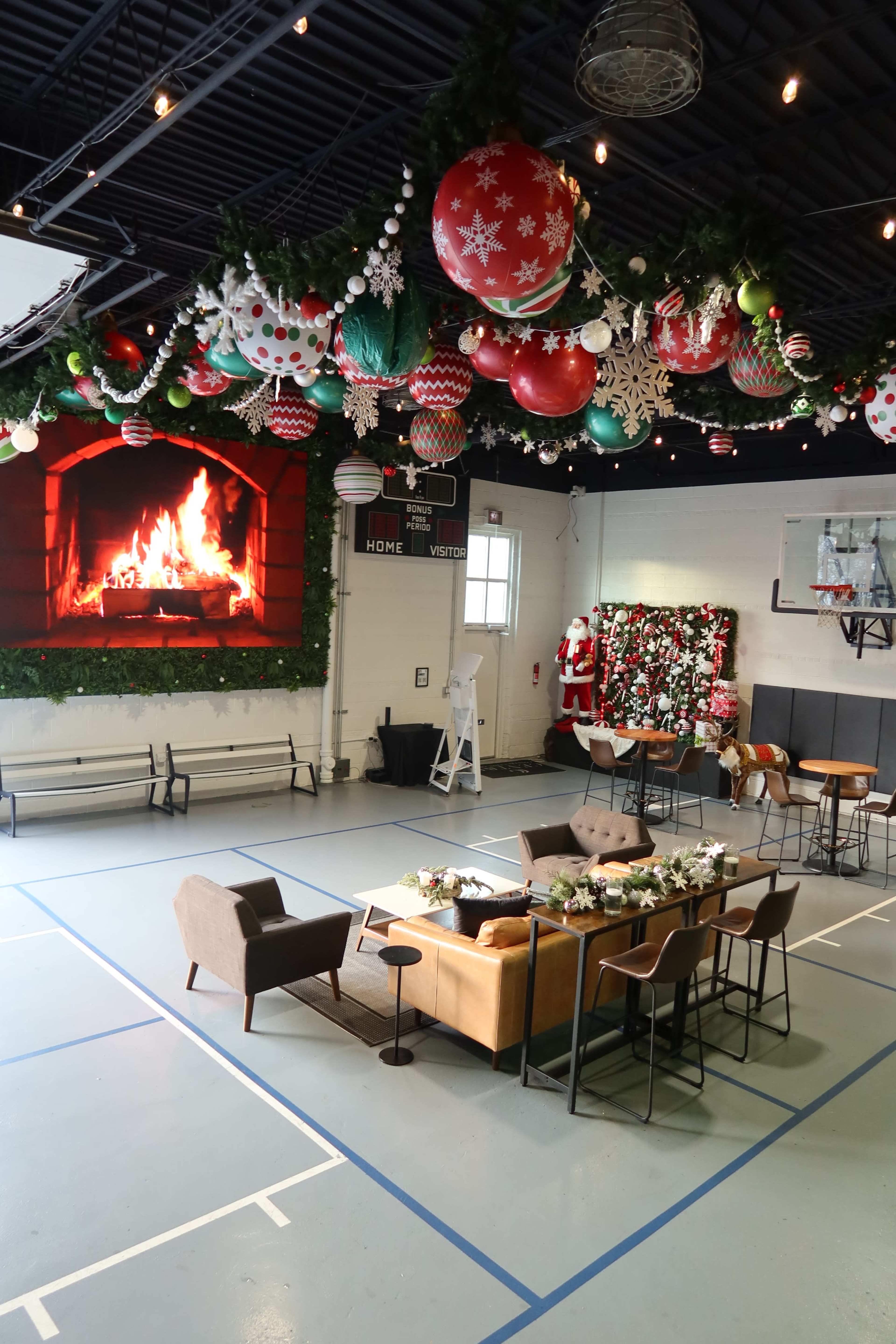 The image shows a decorated indoor space with a faux fireplace, holiday ornaments hanging from the ceiling, and a seating area arranged with furniture for gatherings.