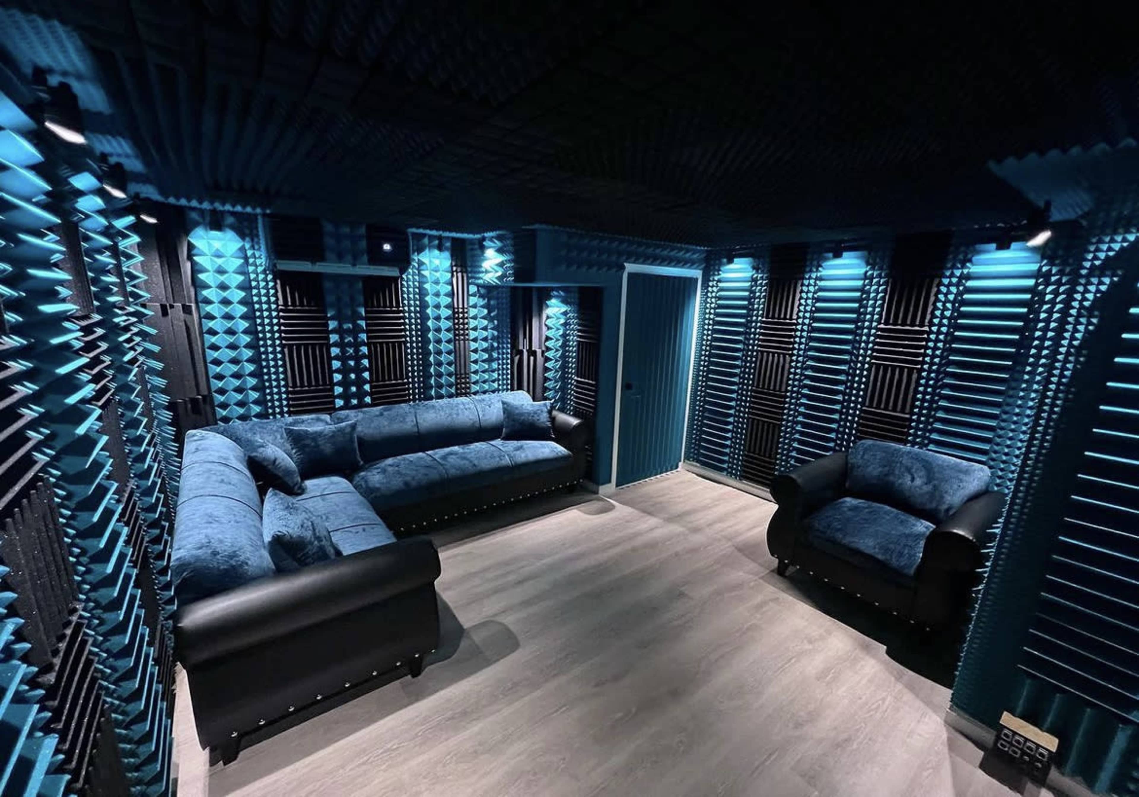 A soundproof room featuring blue acoustic foam panels on the walls, a gray sofa, and a black armchair.