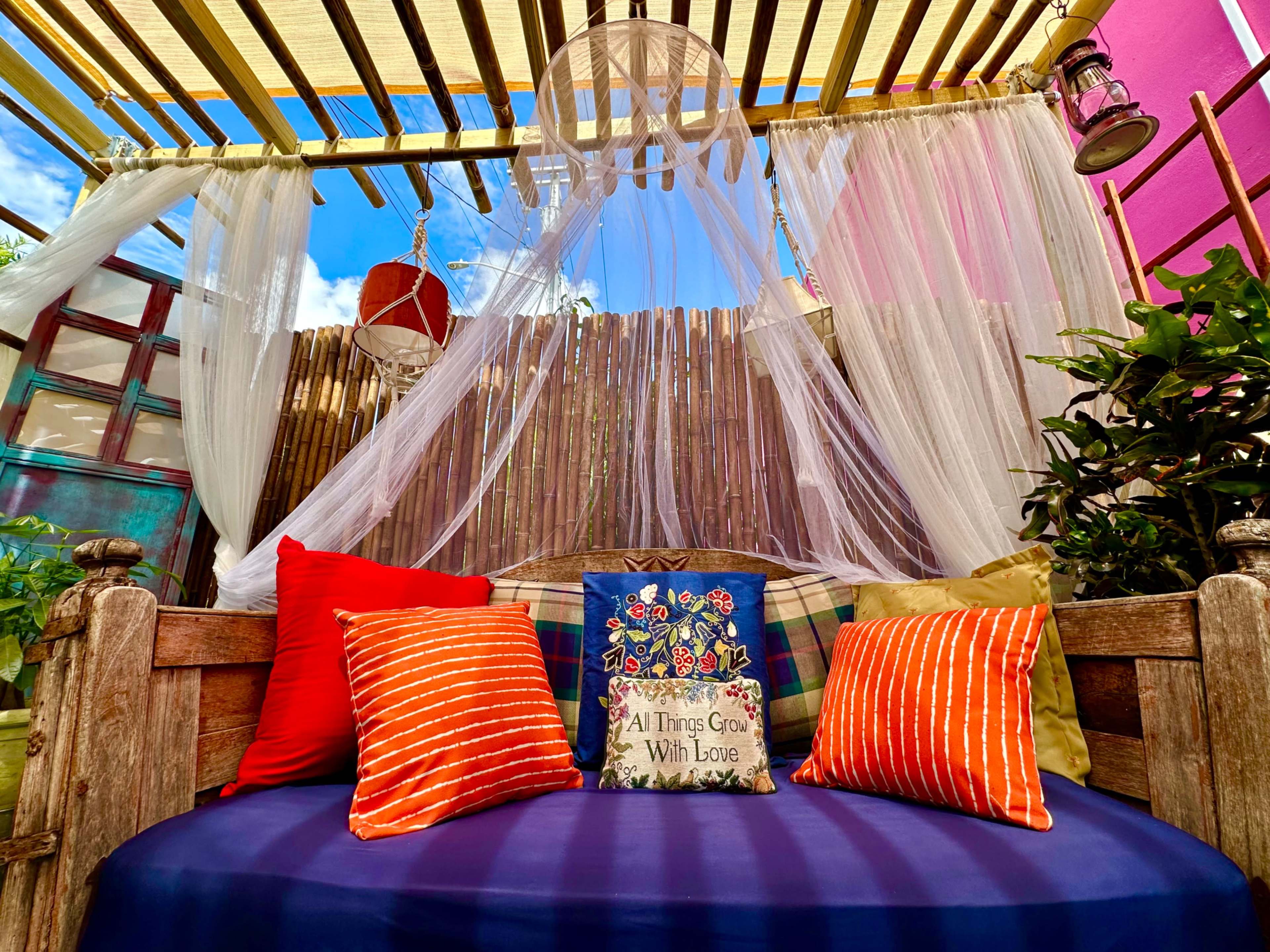 A cozy seating area is decorated with colorful cushions and draped fabrics, set against a backdrop of plants and a wooden fence.
