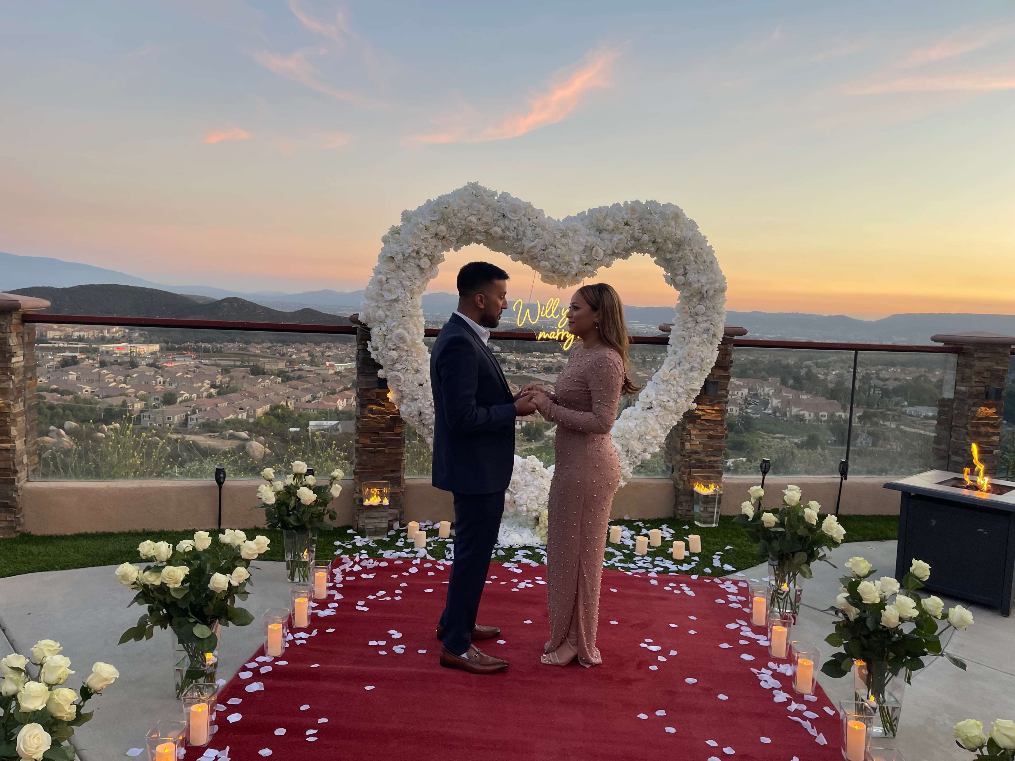 A couple stands holding hands on a red carpet surrounded by white roses and candles in front of a heart-shaped floral arrangement at sunset.