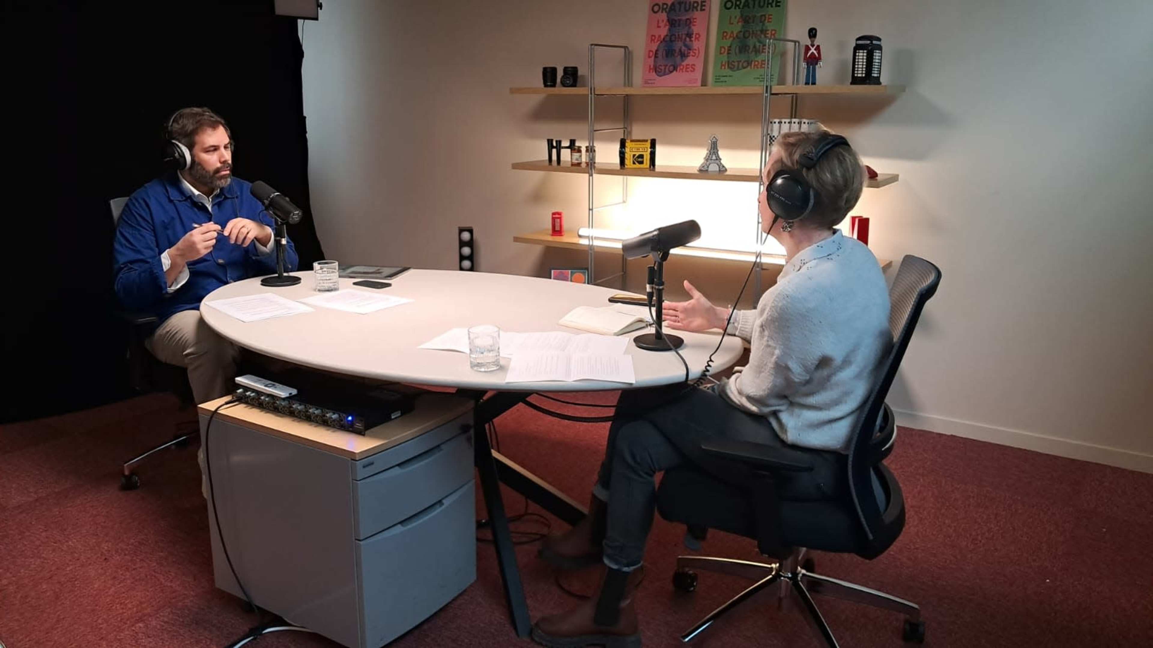 Two people engage in a discussion at a circular table in a studio setting equipped with microphones and various decor items on the shelves.