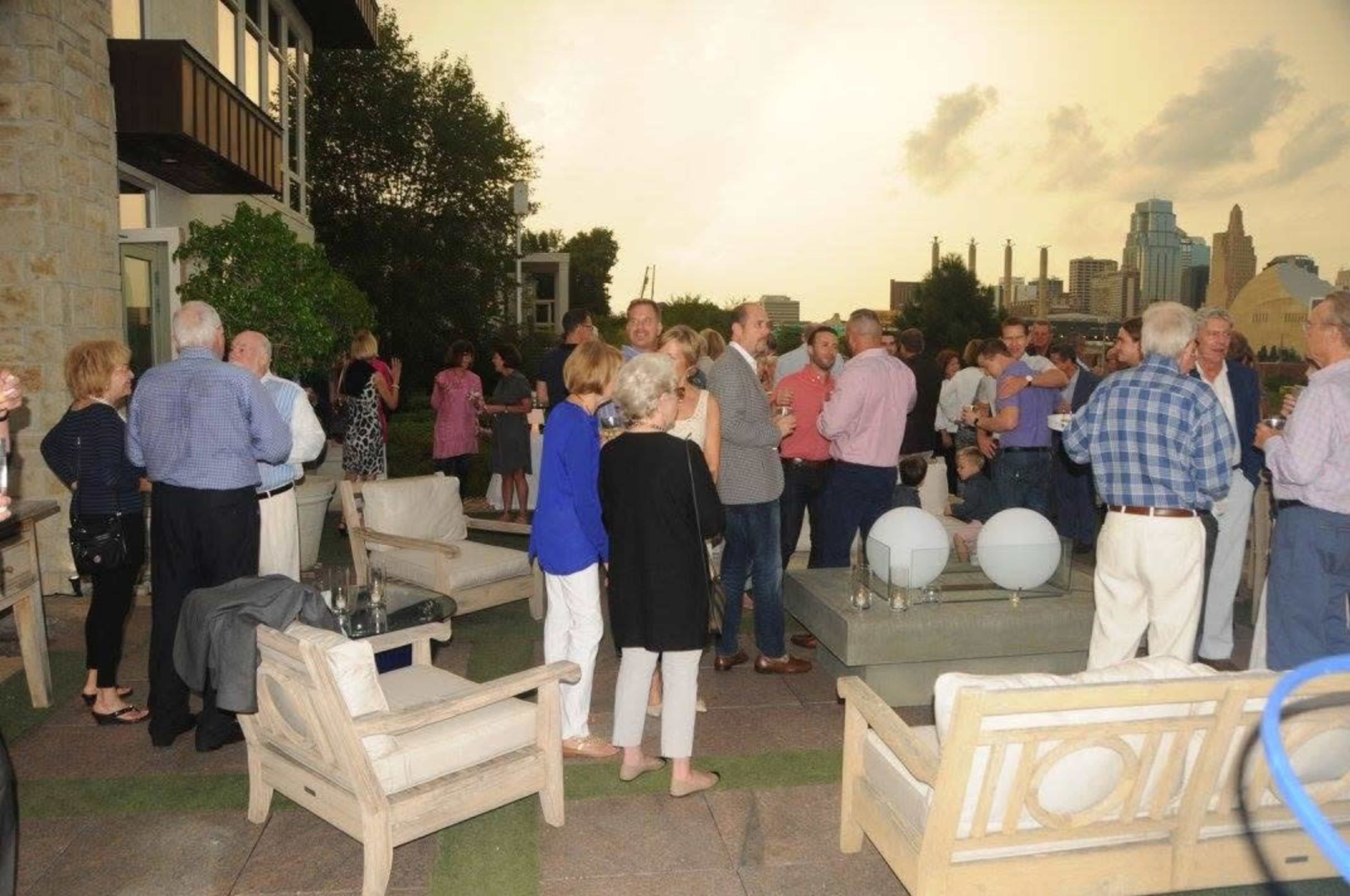 A large group of people mingles on a rooftop terrace with a city skyline in the background during an evening gathering.
