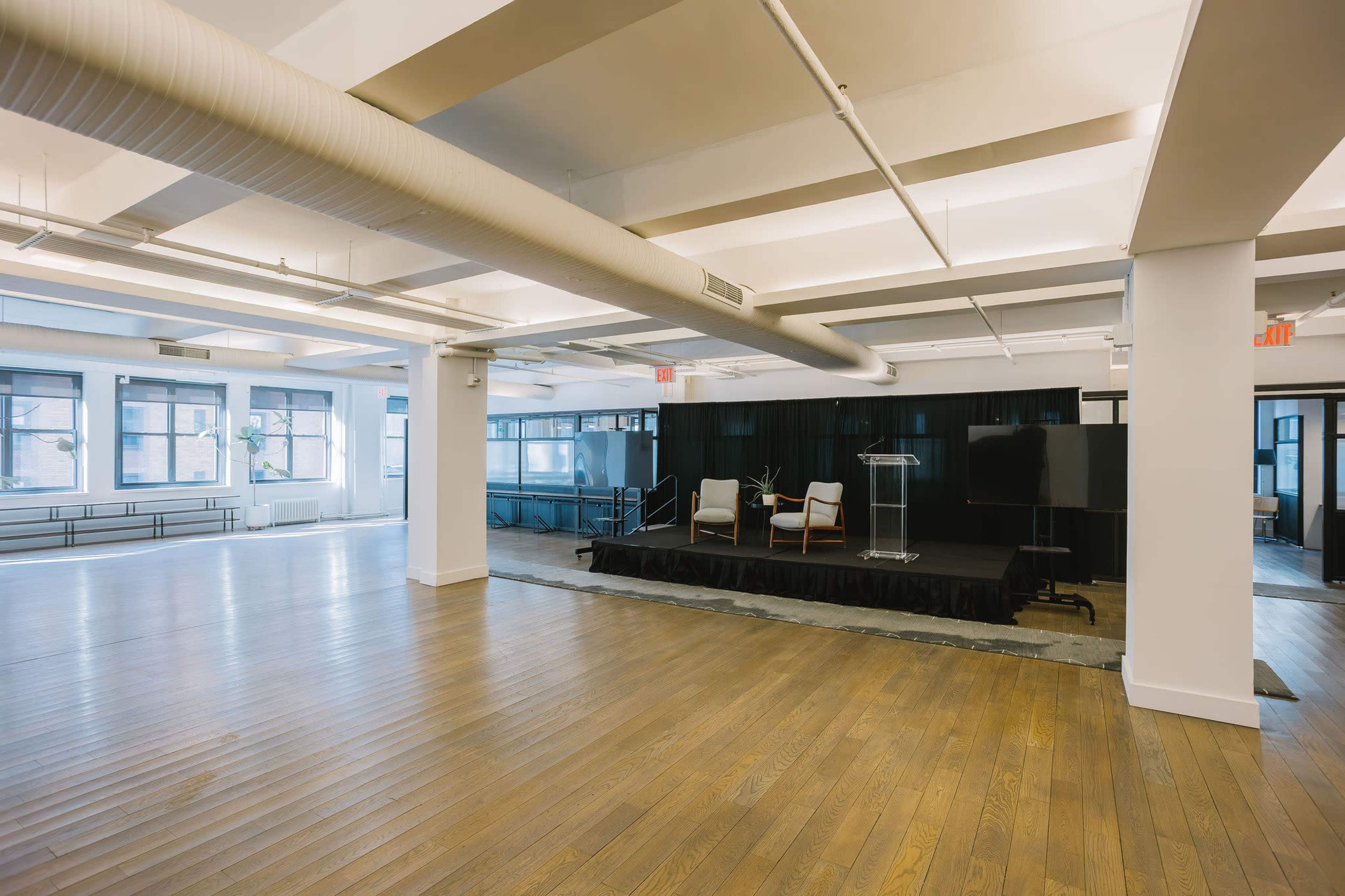 The image shows a large, empty event space with a stage at one end, featuring two chairs and a podium, surrounded by large windows.