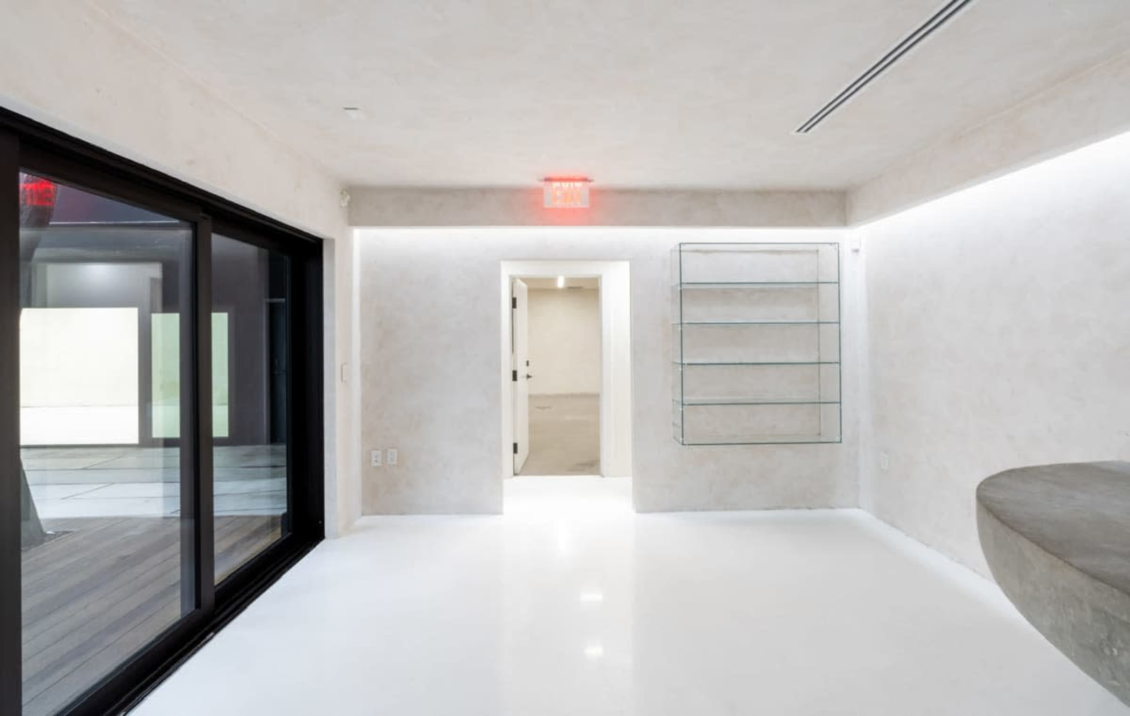 The image shows a minimalist indoor space with smooth white walls, a polished floor, and a glass shelving unit against one wall.