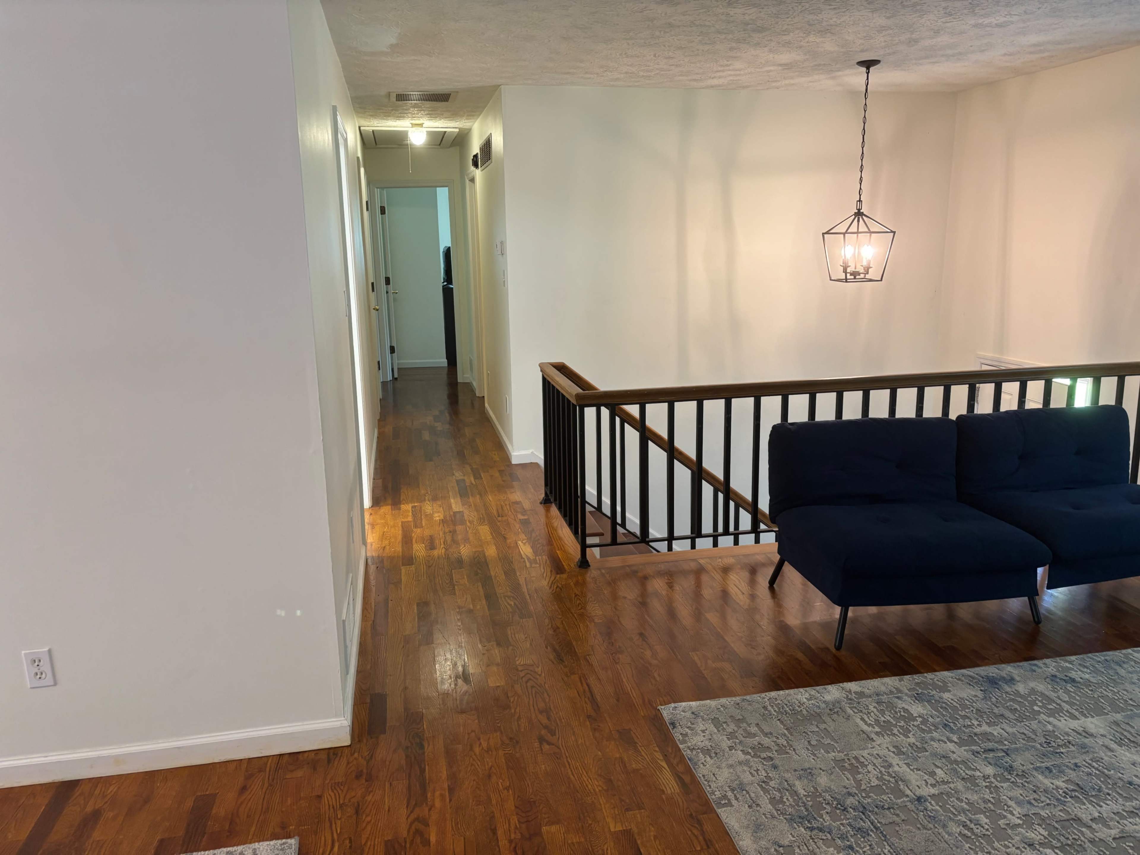 The image shows a well-lit interior space featuring a wooden floor, a dark blue sofa, and a hallway leading to a door in the background.