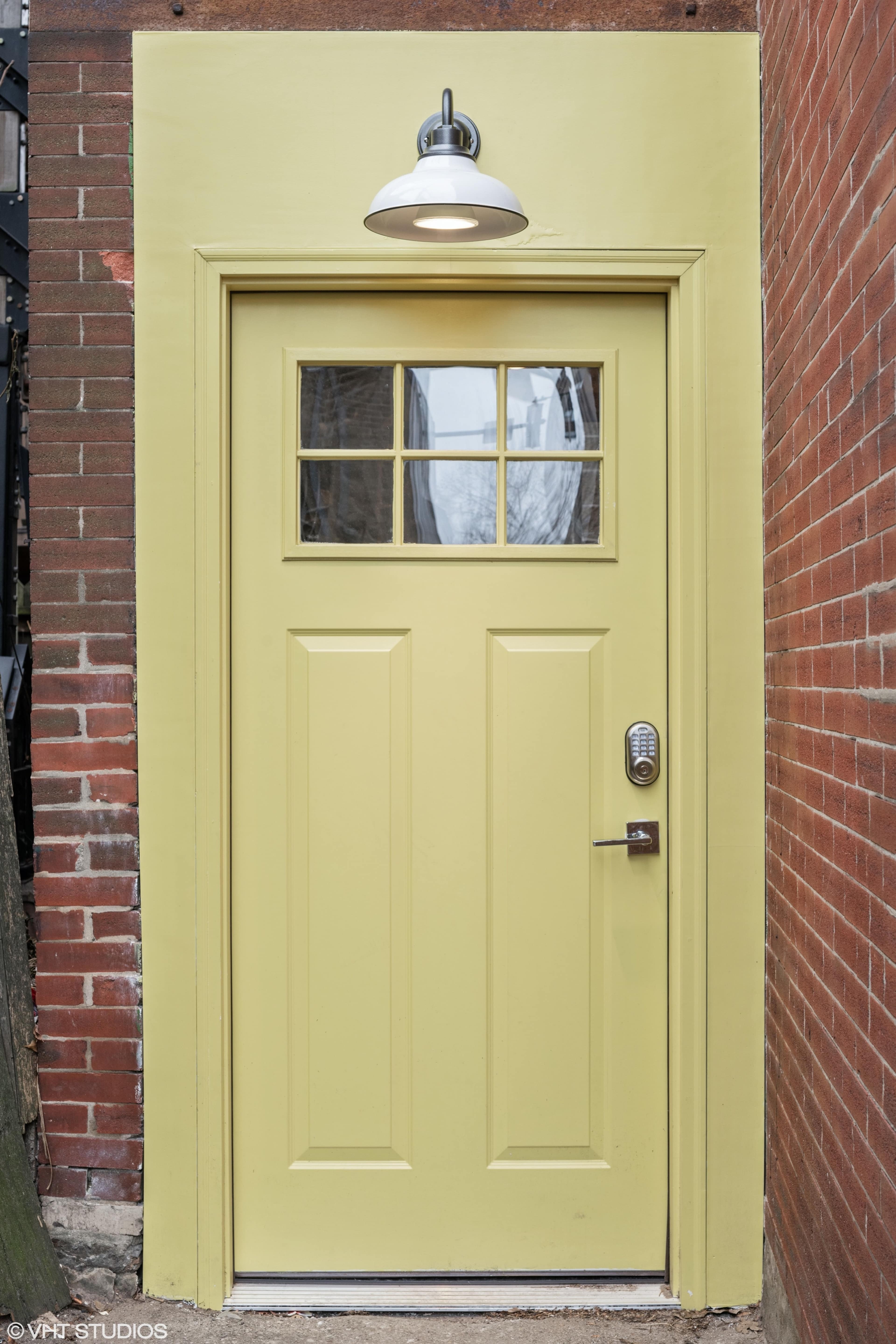 A yellow door with a small window at the top, set in a brick wall and illuminated by a wall-mounted light fixture.