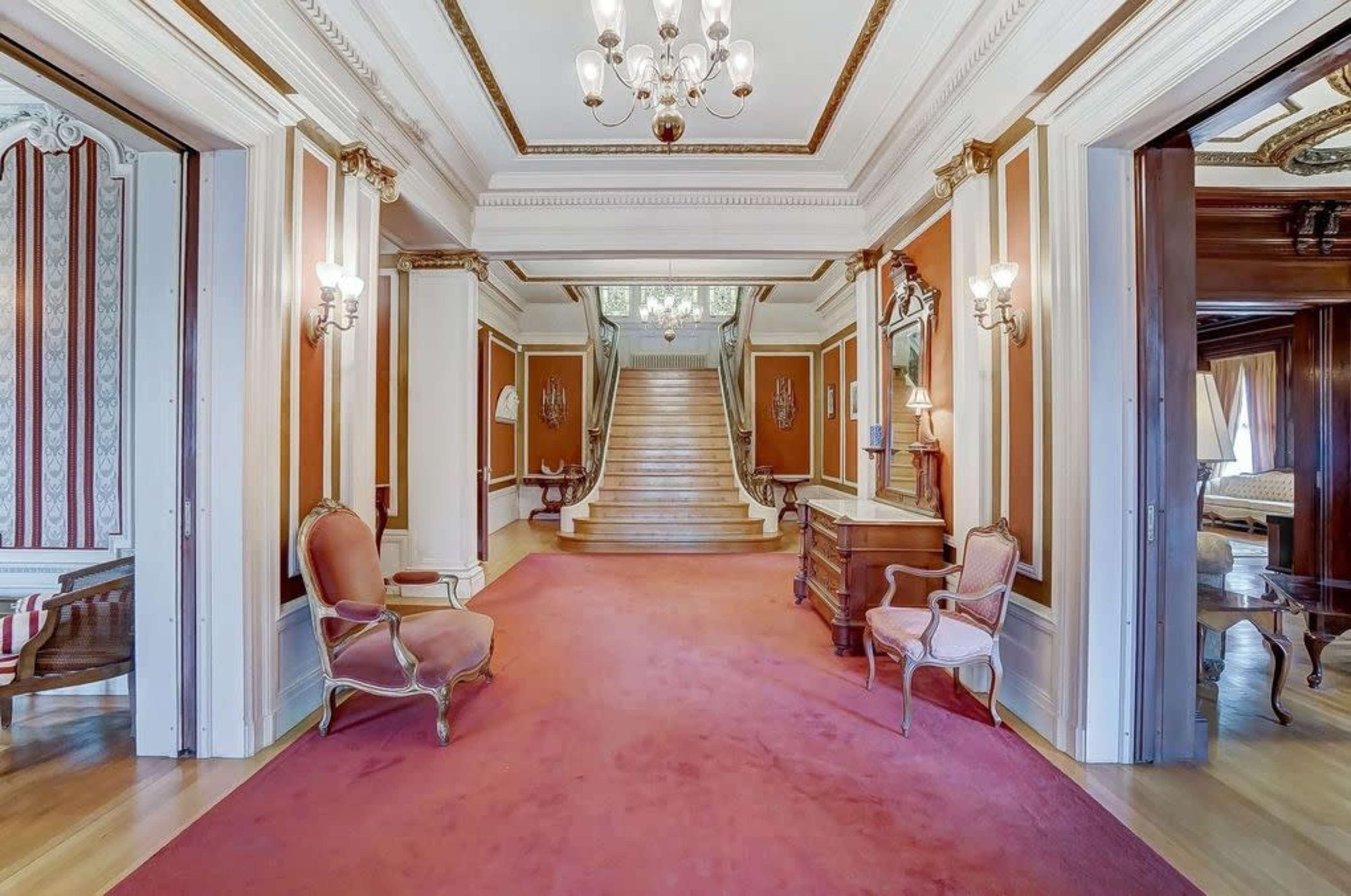 An elegant hallway with a patterned carpet, leading to a staircase, flanked by ornate lighting and decorative wall panels.