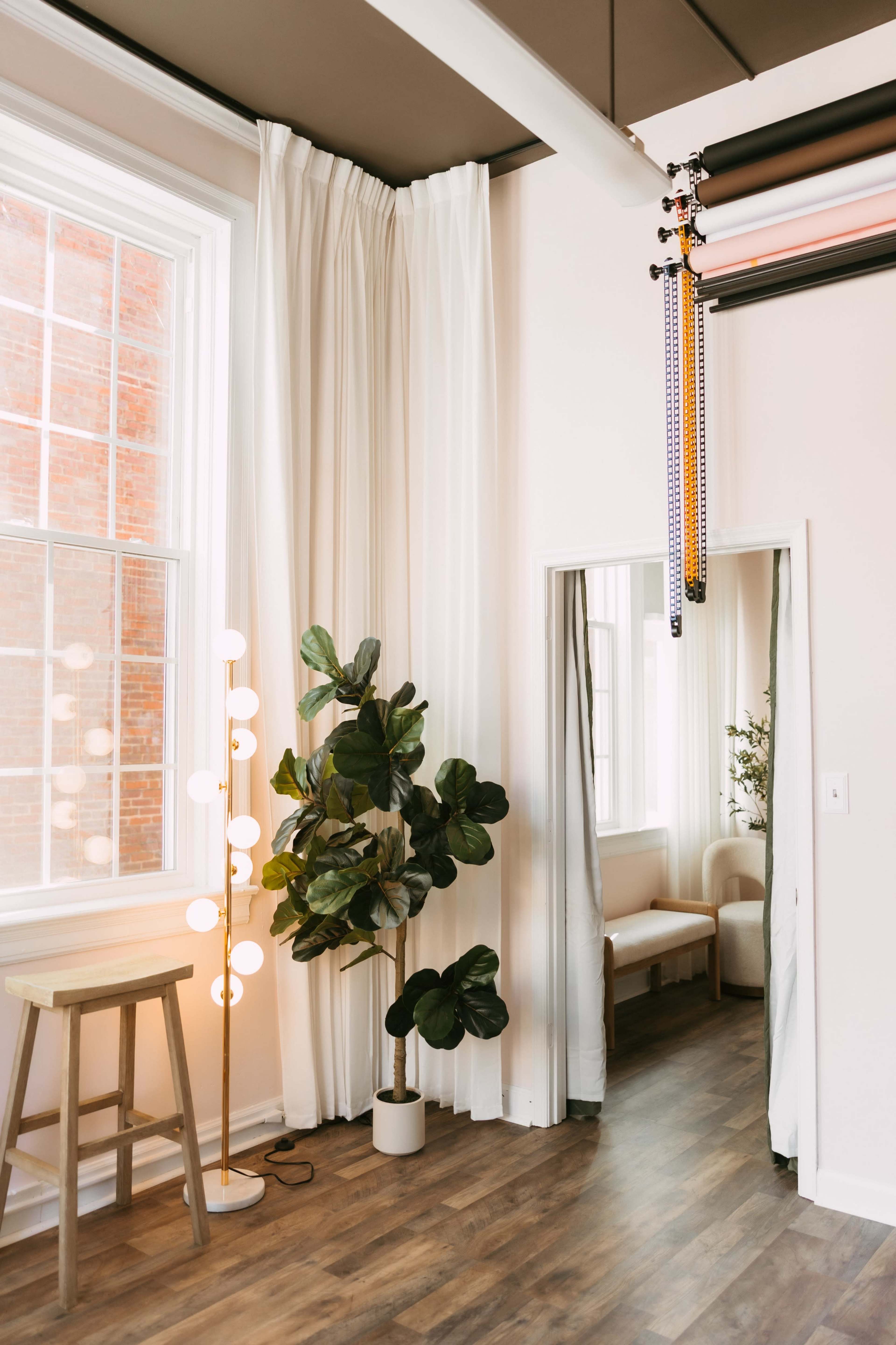 The image shows a bright room featuring a window with white curtains, a modern floor lamp, a potted plant, a wooden stool, and a mirror reflecting a seating area.