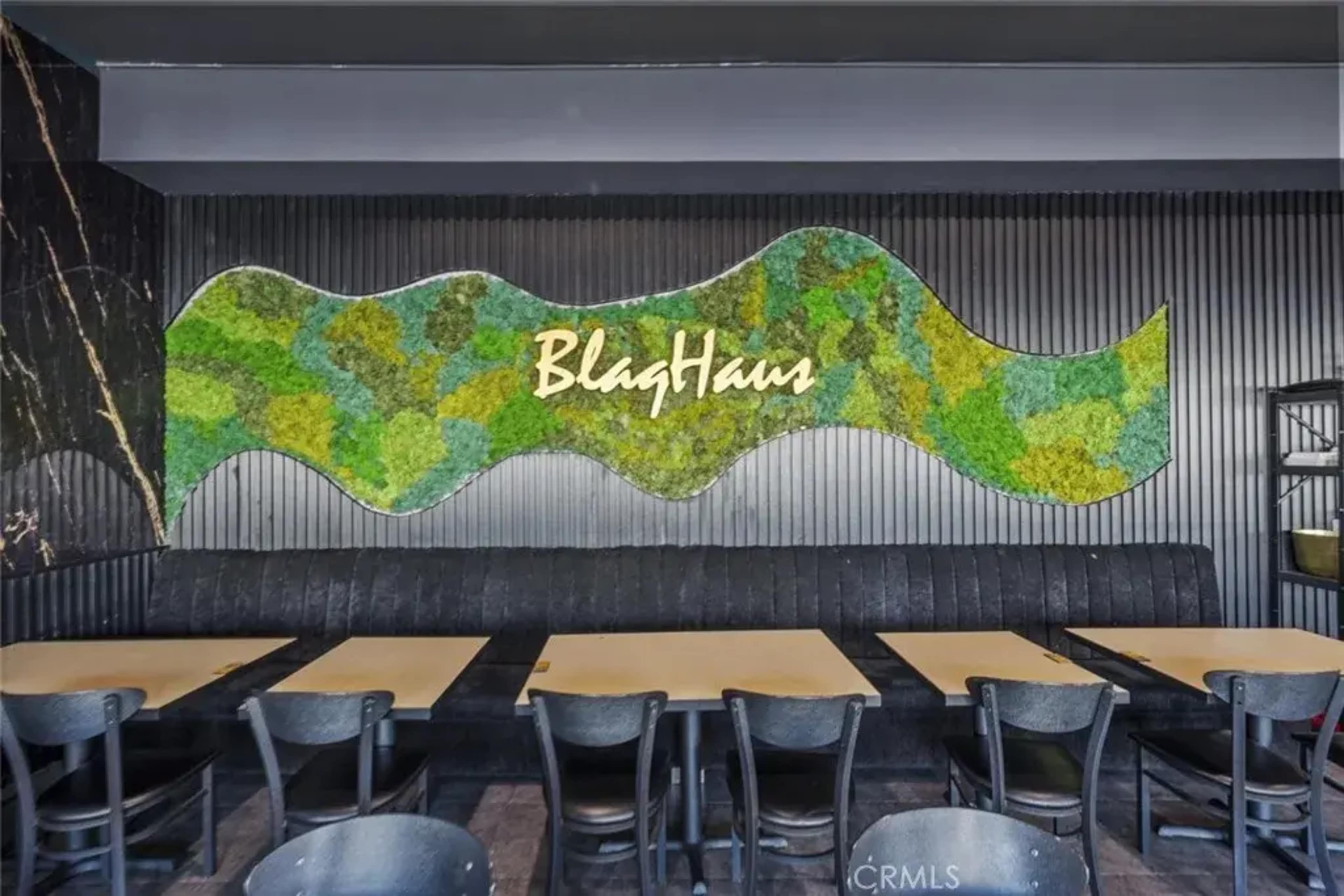 The image shows the interior of a restaurant featuring a moss wall art piece with the name "BlagHaus" above a black upholstered bench and wooden tables arranged in front.