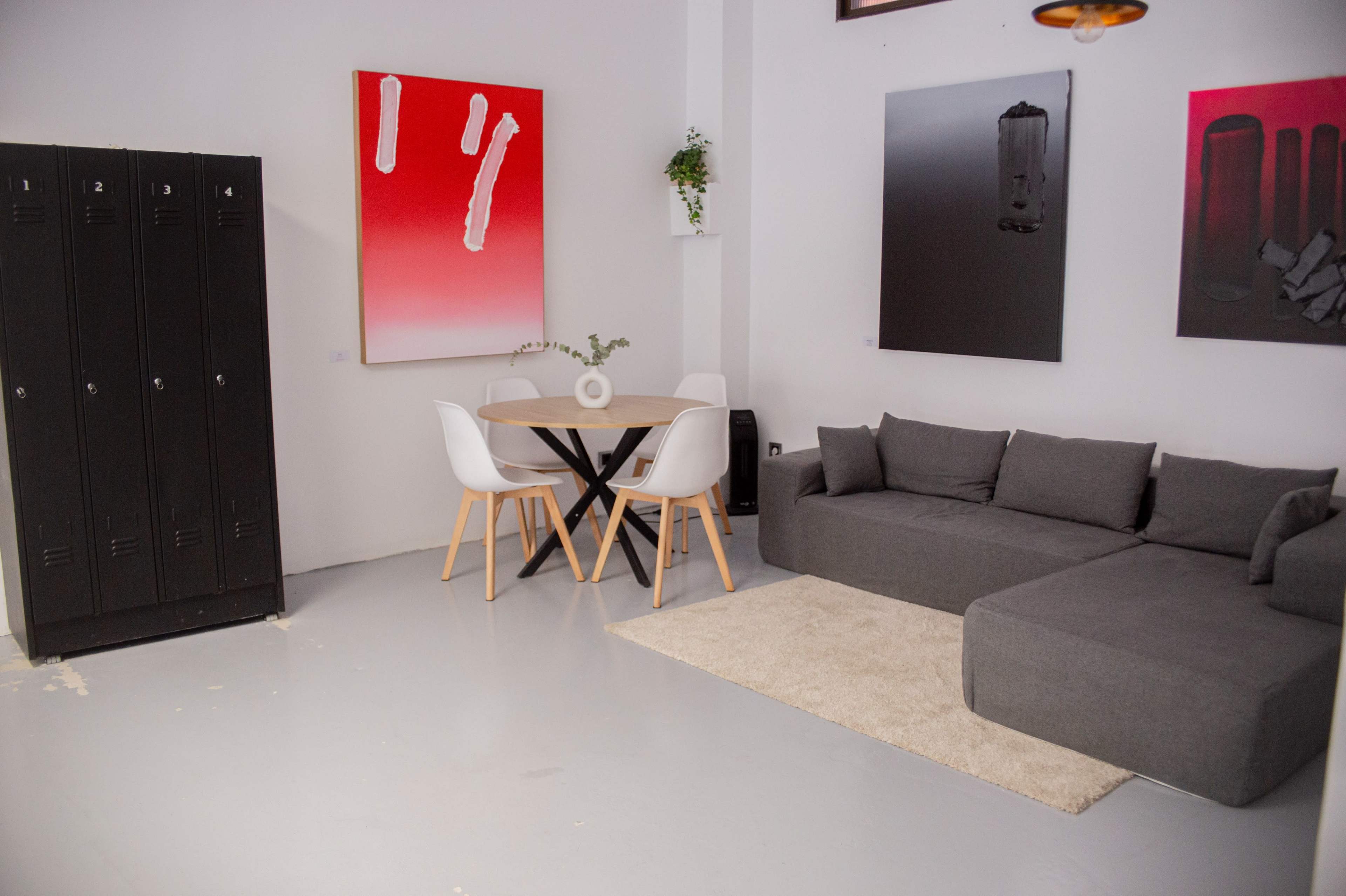 The image shows a minimalist living space with a gray sofa, a round dining table surrounded by white chairs, and black lockers against the wall, complemented by abstract wall art.