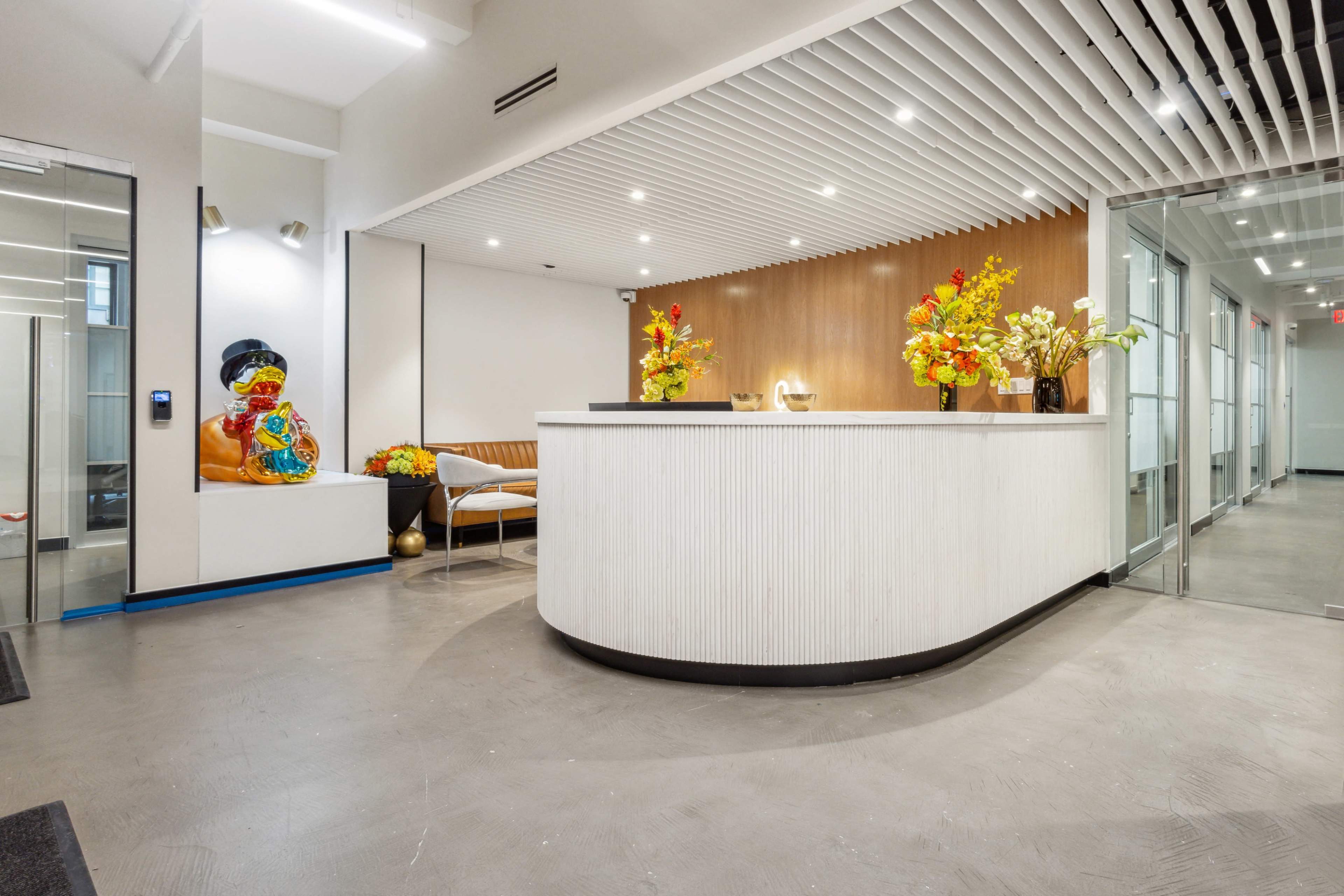 The image shows a modern reception area featuring a curved front desk, decorative flower arrangements, and a colorful statue against the wall.