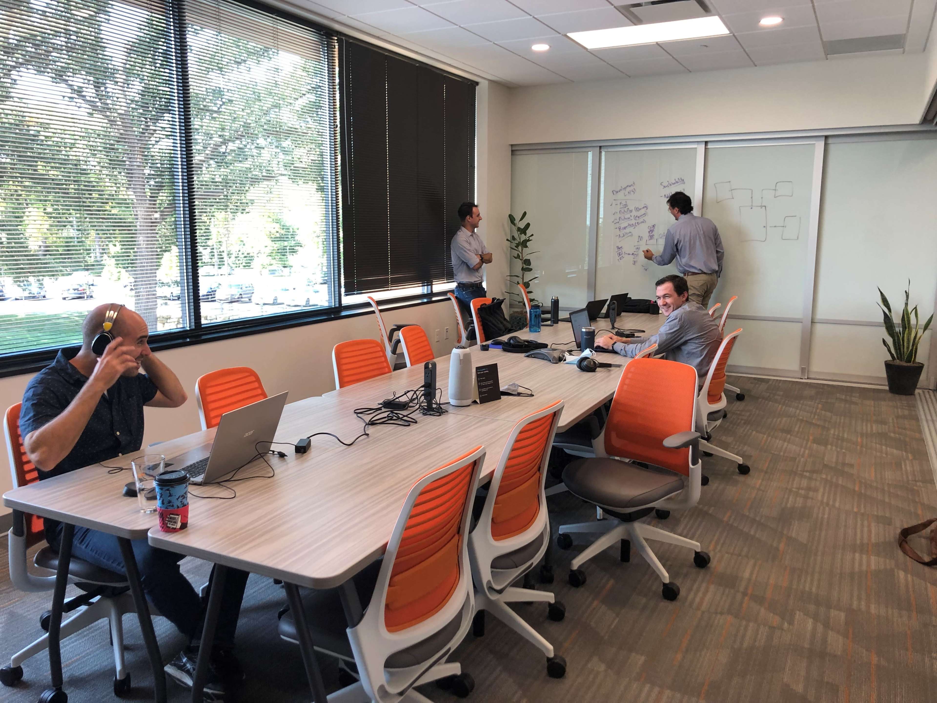 Four people are working in a conference room, with two seated at a table using laptops and two standing near a whiteboard.
