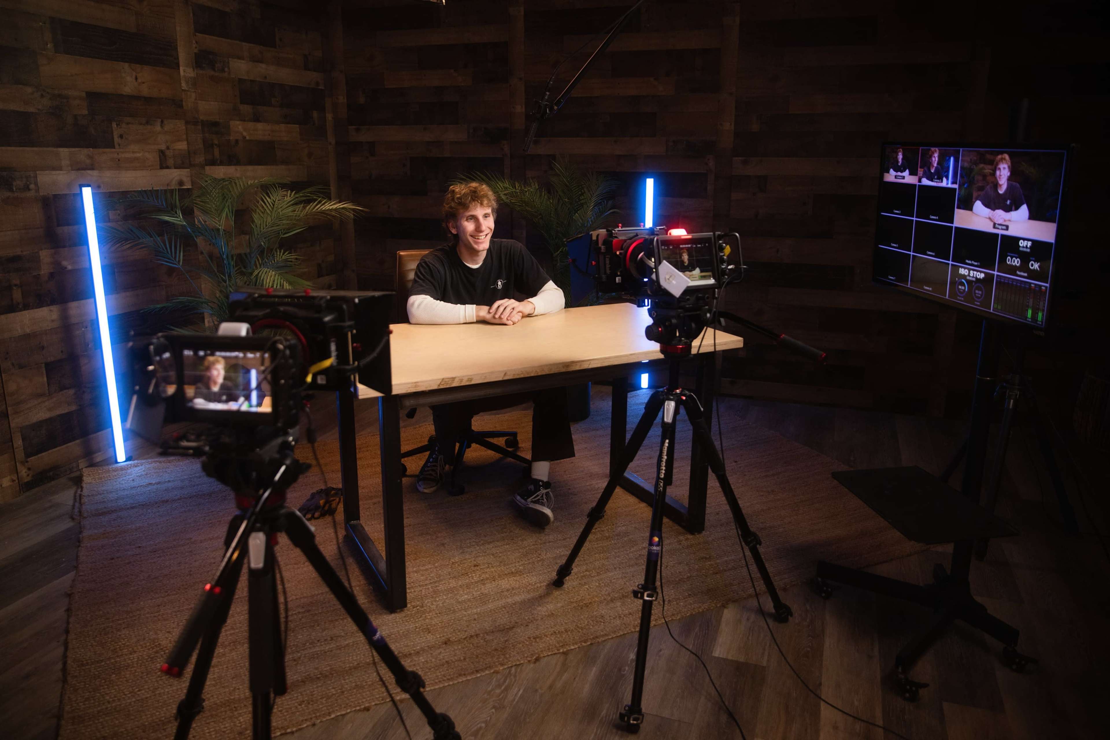 A person sits at a wooden table in a studio setup surrounded by cameras and a monitor displaying a video feed.
